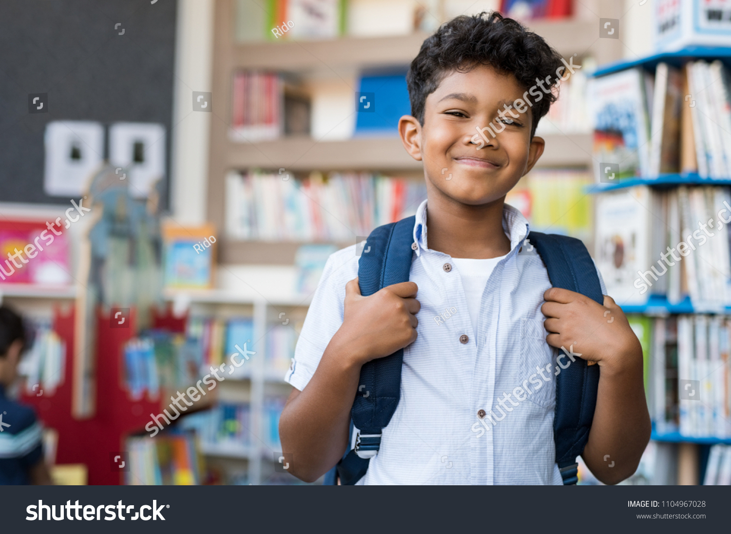 Portrait of smiling hispanic boy looking at camera. Young elementary schoolboy carrying backpack and standing in library at school. Cheerful middle eastern child standing with library background. #1104967028