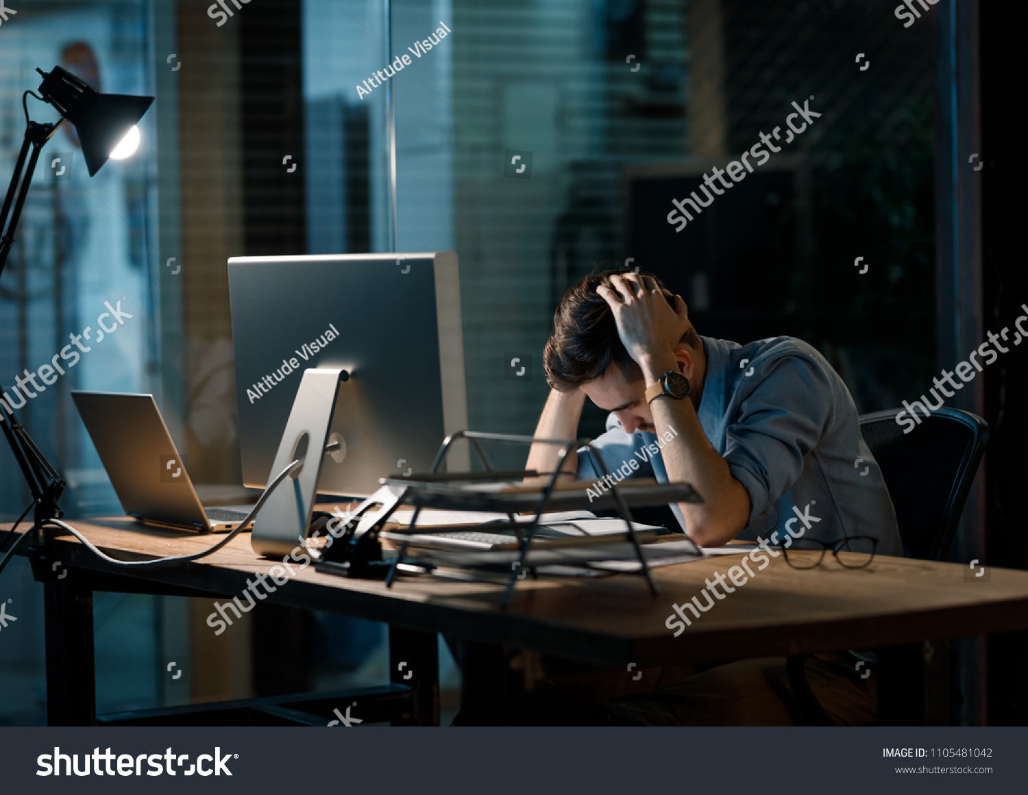 Young man in shirt working alone in office late sitting in lamplight at table and looking sleepy.  #1105481042