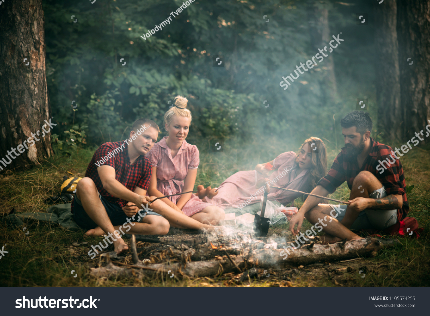 Two couples having picnic in woods. Bearded man and his best friend cooking sausages over fire. Young hikers around campfire in the evening. Friendship and leisure concept. #1105574255