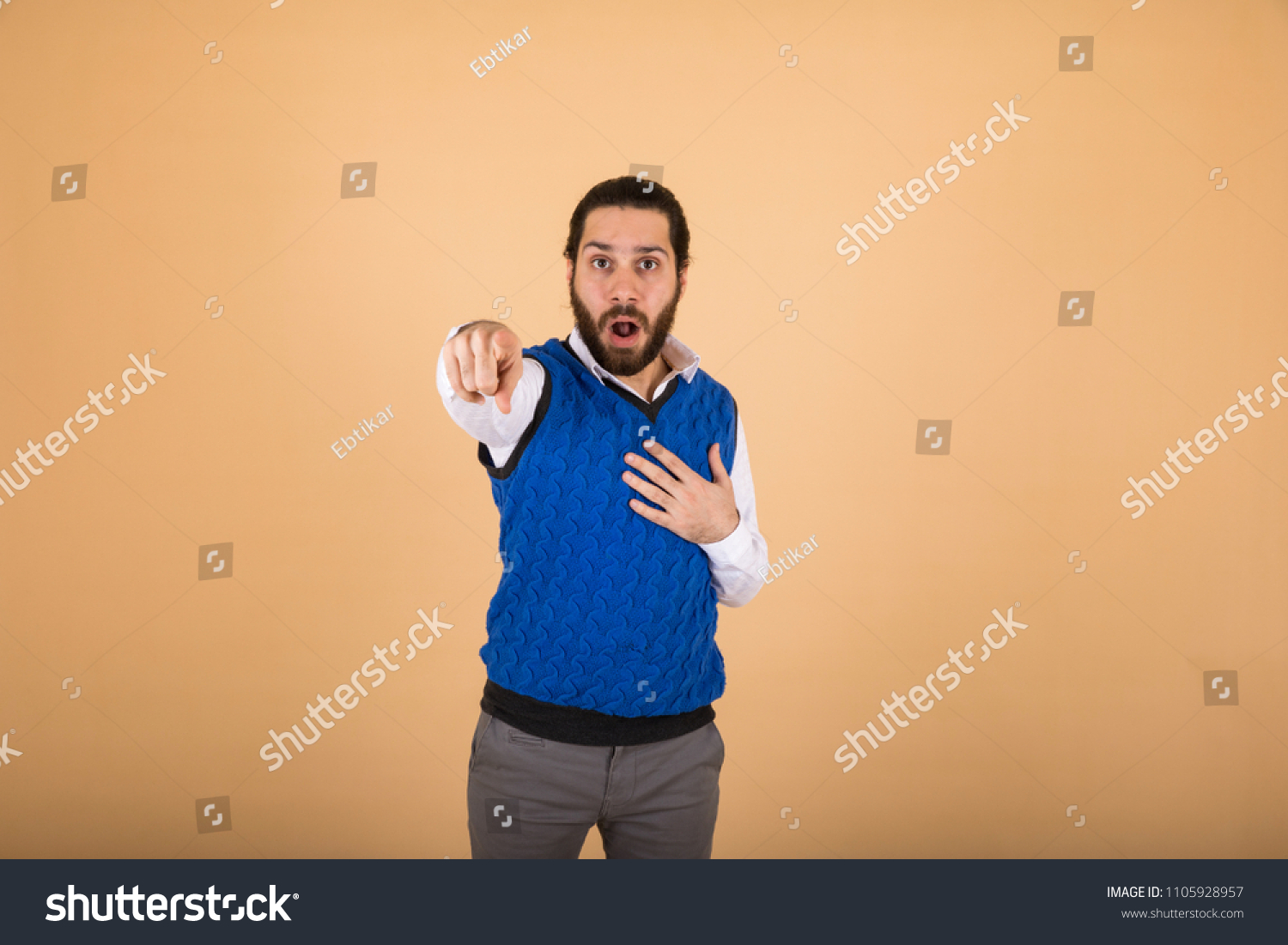 young man shocked and pointing at something and looking to the camera , in an orange background #1105928957