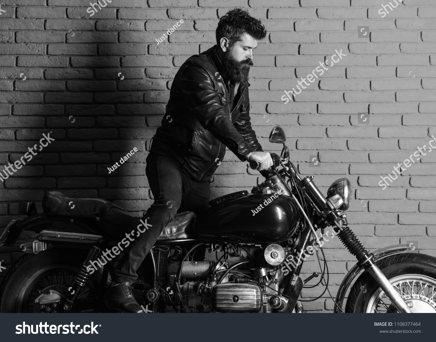 man on a motorcycle. Hipster, brutal biker on serious face in leather jacket gets on motorcycle. Man with beard, biker in leather jacket near motor bike in garage, brick wall background #1108377464