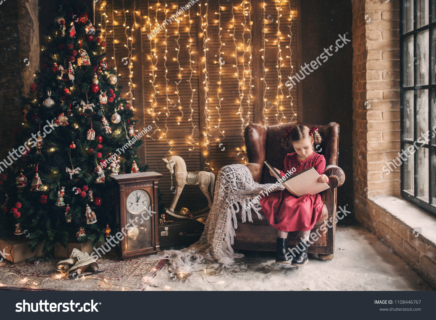 little girl is sitting in a chair and thumbs through a vintage photo album next to a Christmas tree on a background of Christmas lights, photo in retro style #1108446767