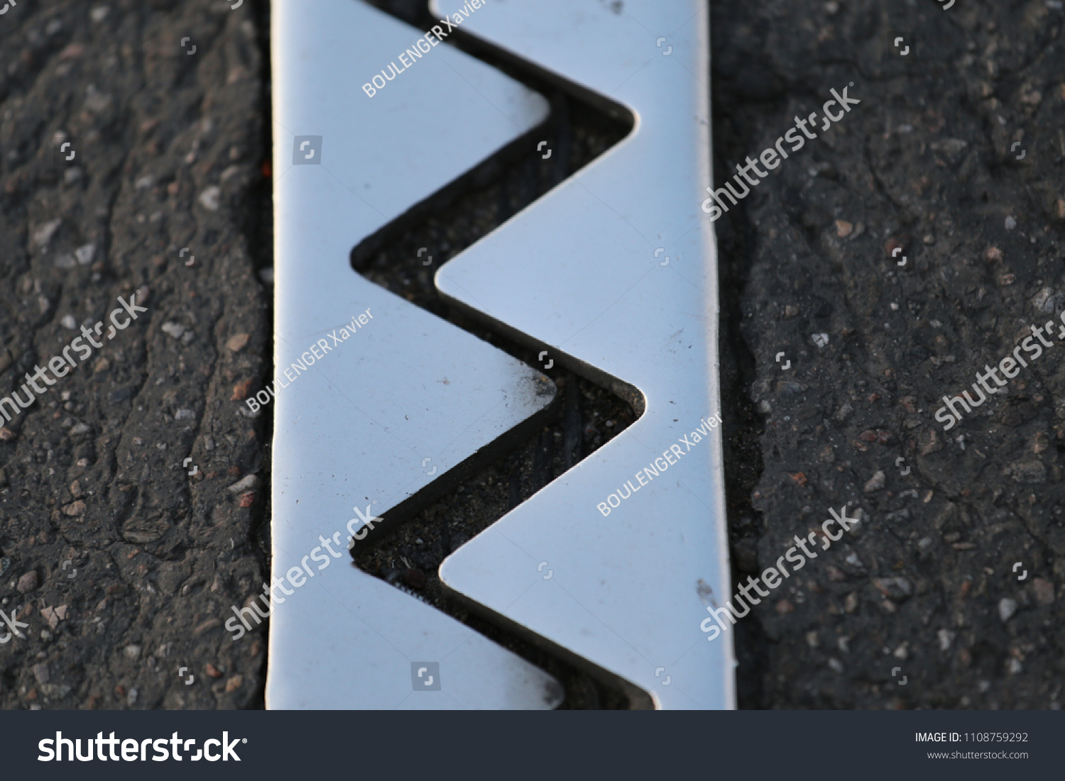 Close up outdoor view from above of an iron dilatation joint placed on a bridge road. Zigzag line drawn of an bright steel surface. Abstract design with grey geometric shapes on the asphalt ground.   #1108759292