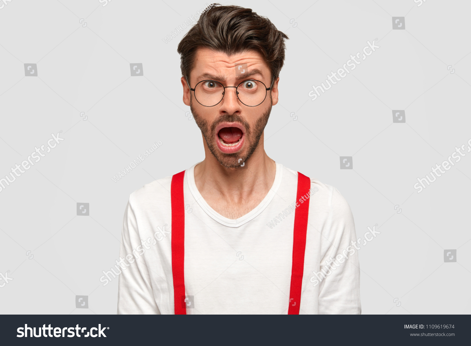 Puzzled emotive bearded male reacts on something unexpected, frowns face and exclaims with stupefaction, wears fashionable shirt and red braces, isolated on white background. People, emotions concept #1109619674