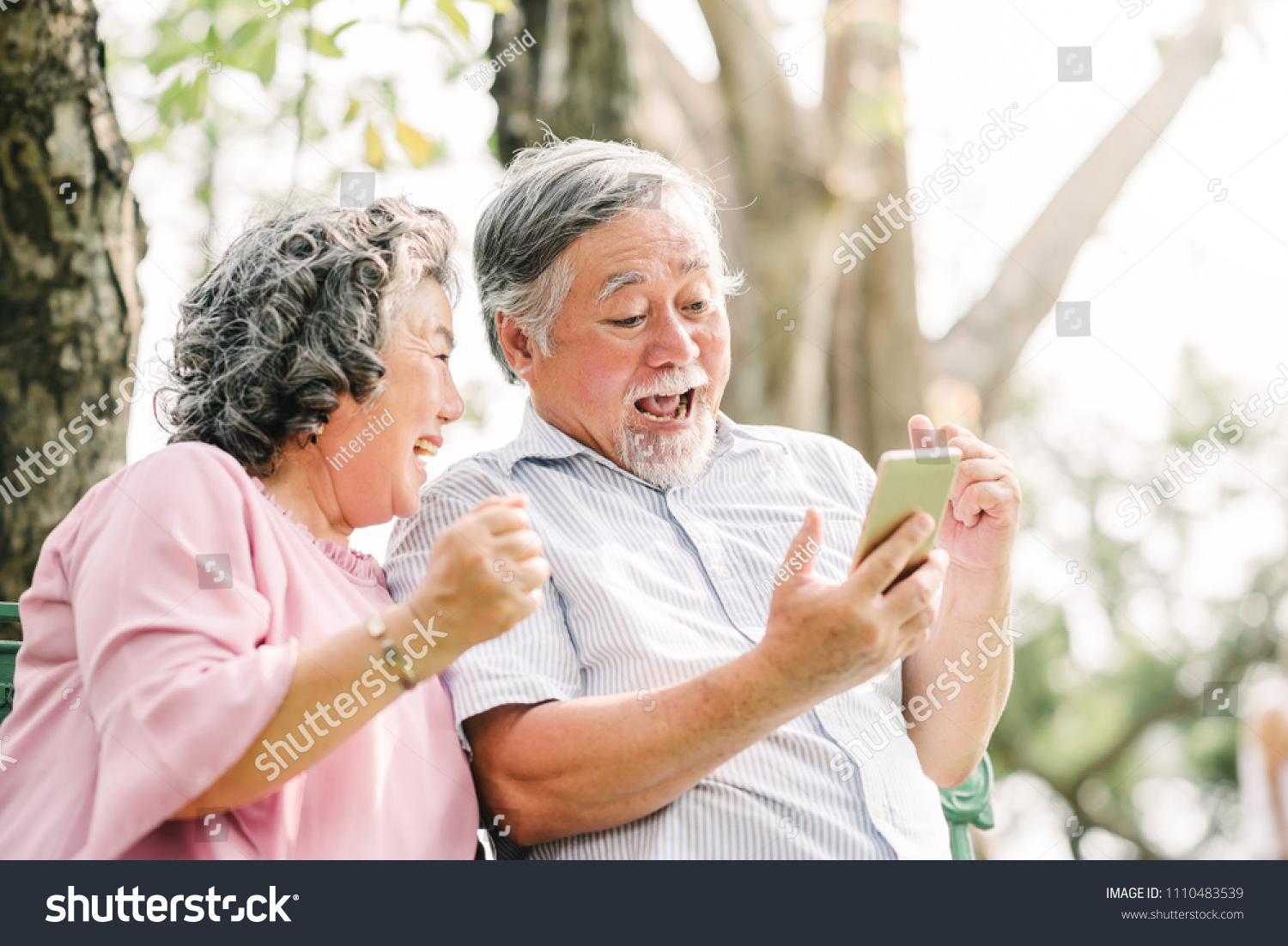 Happy senior Asian couple laughing and exciting with something in smartphone mobile while sitting in the park #1110483539
