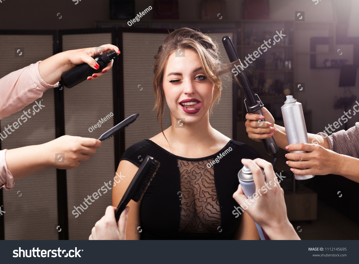 beautiful emotional woman is surrounded by many hands holding hair curler, sprays and brushes. girl is showing a tongue with one eye closed. concept of professional makeup products #1112145695