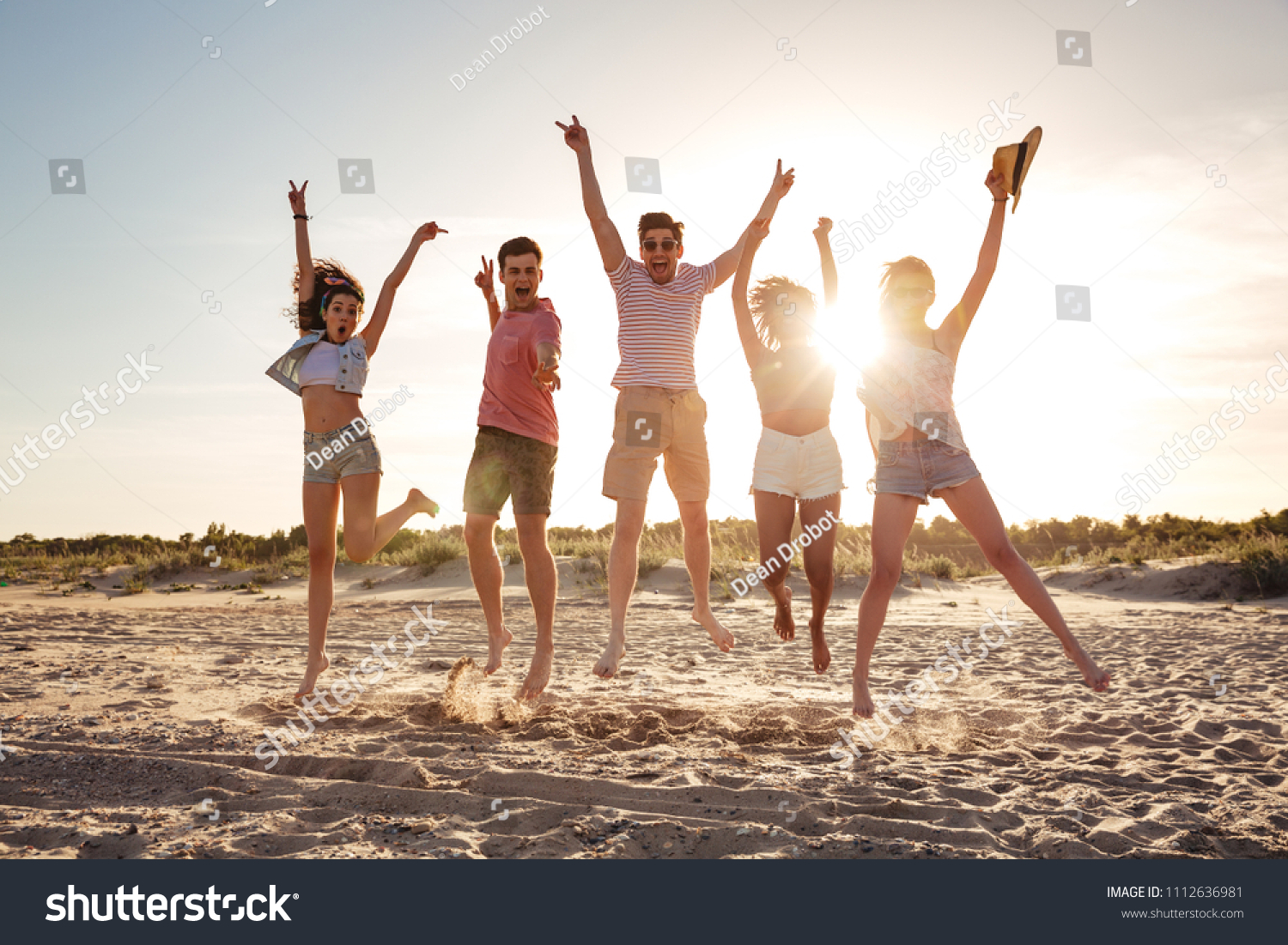 Group of cheerful young friends dressed in summer clothes jumping together with hands raised at he beach #1112636981