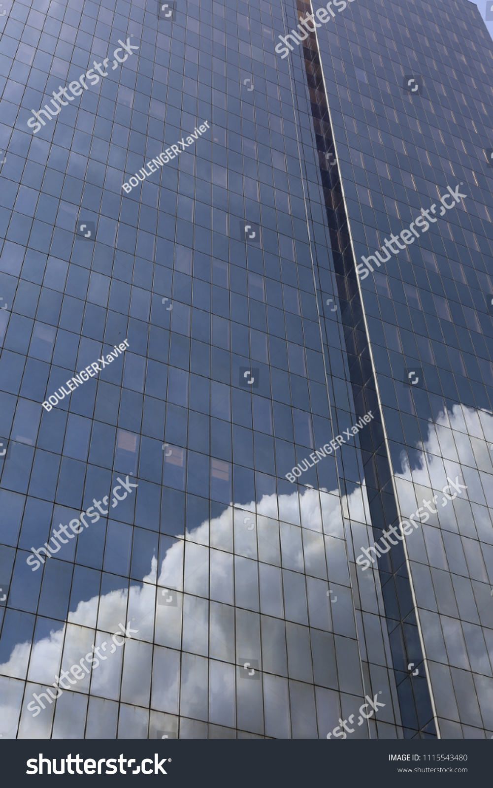 Close up outdoor view of part of a skyscraper with pattern of reflective glass windows. Blue cloudy sky reflected on the bright surface. Modern architecture with a big white cloud on the facade.  #1115543480