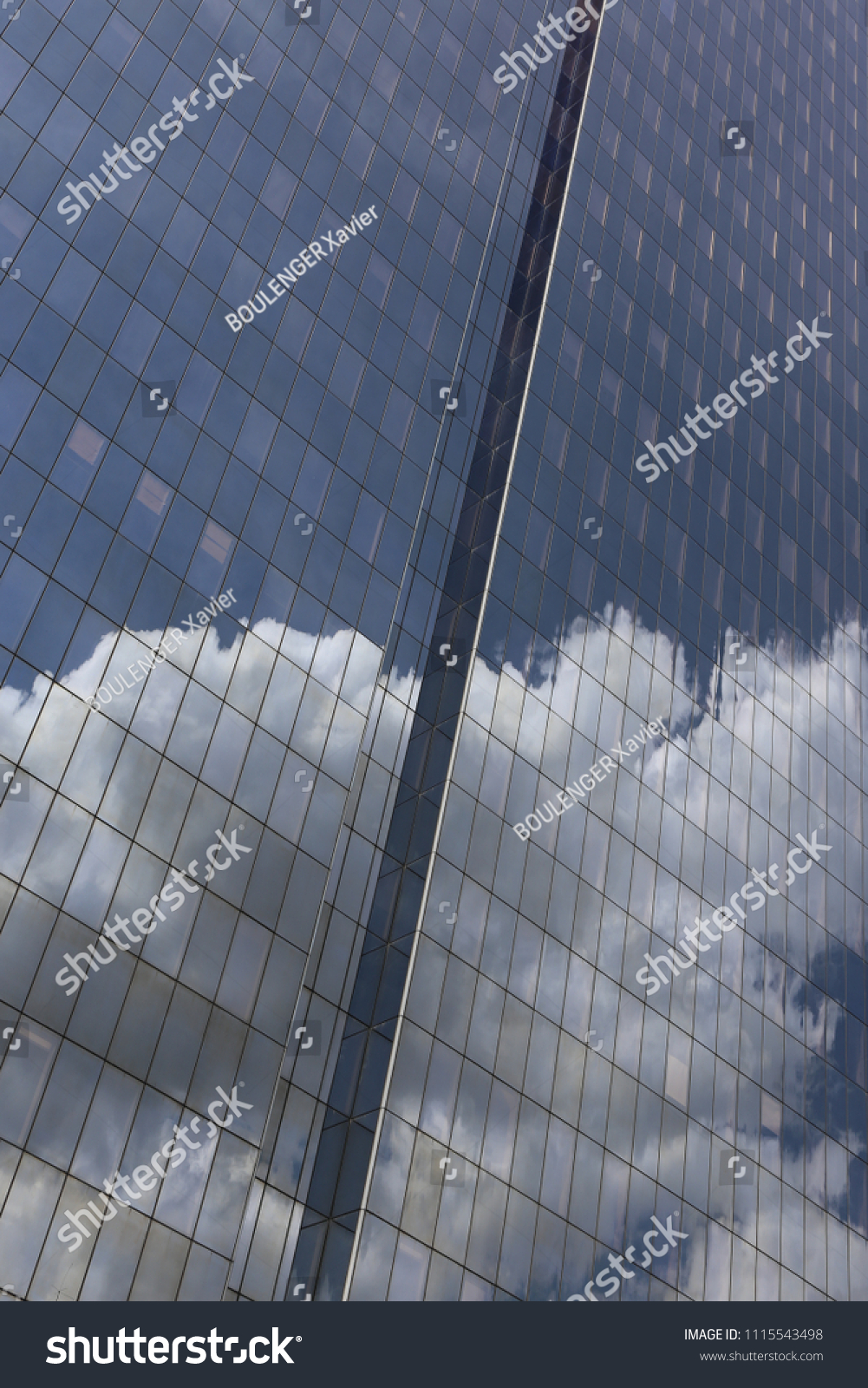 Close up outdoor view of part of a skyscraper with pattern of reflective glass windows. Blue cloudy sky reflected on the bright surface. Modern architecture with a big white cloud on the facade.  #1115543498