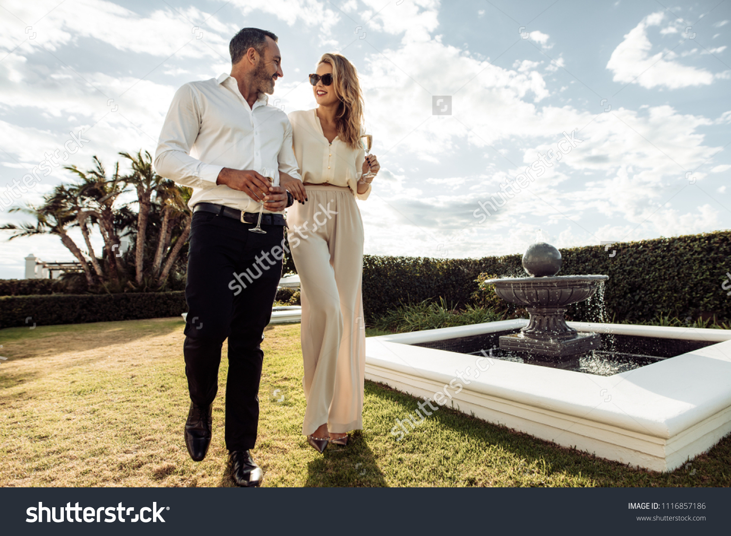 Stylish couple walking outdoors in lawn with a glass of wine. Man and woman looking at each other and walking together outdoors.. #1116857186