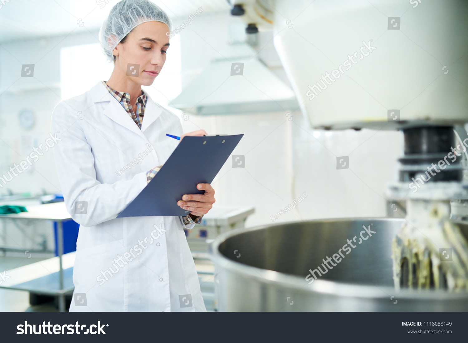 Confectionery factory employee standing in white coat near operating machinery and making notes.  #1118088149