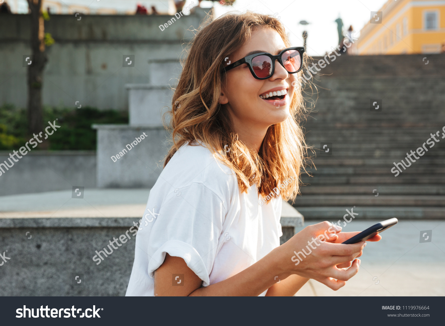 Picture of european brunette woman wearing casual summer outfit and sunglasses laughing while walking through city street with smartphone in hands #1119976664
