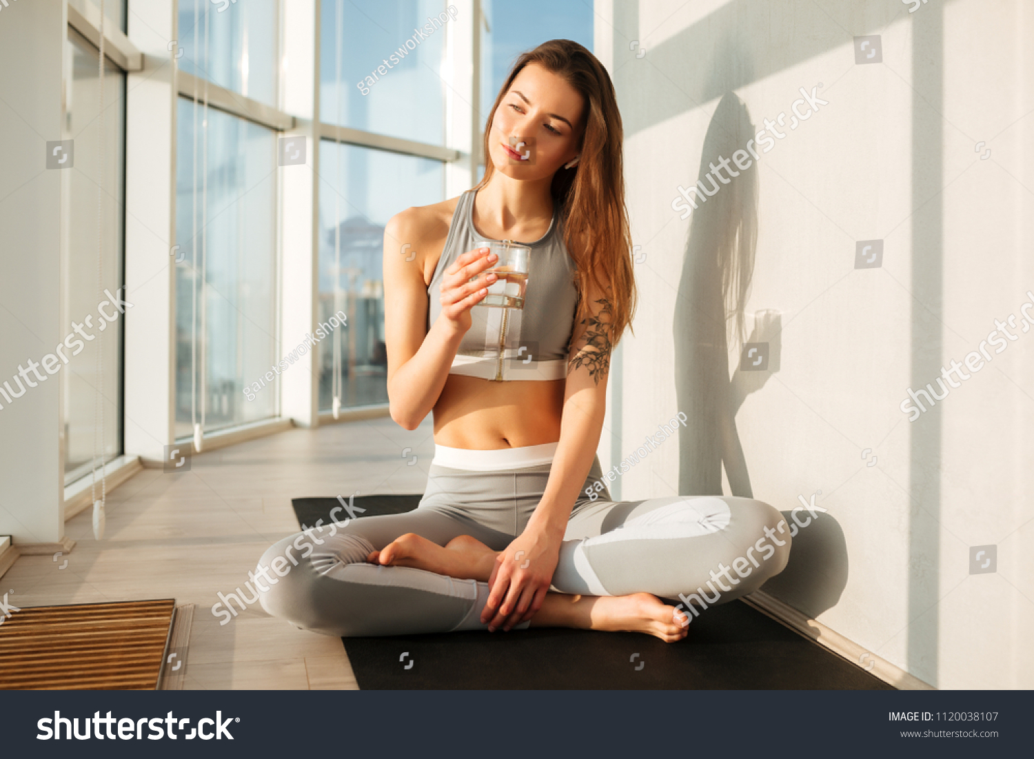 Portrait of thoughtful lady in sporty top and leggings sitting in lotus pose on yoga mat and drinking pure water at home with big beautiful windows on background #1120038107