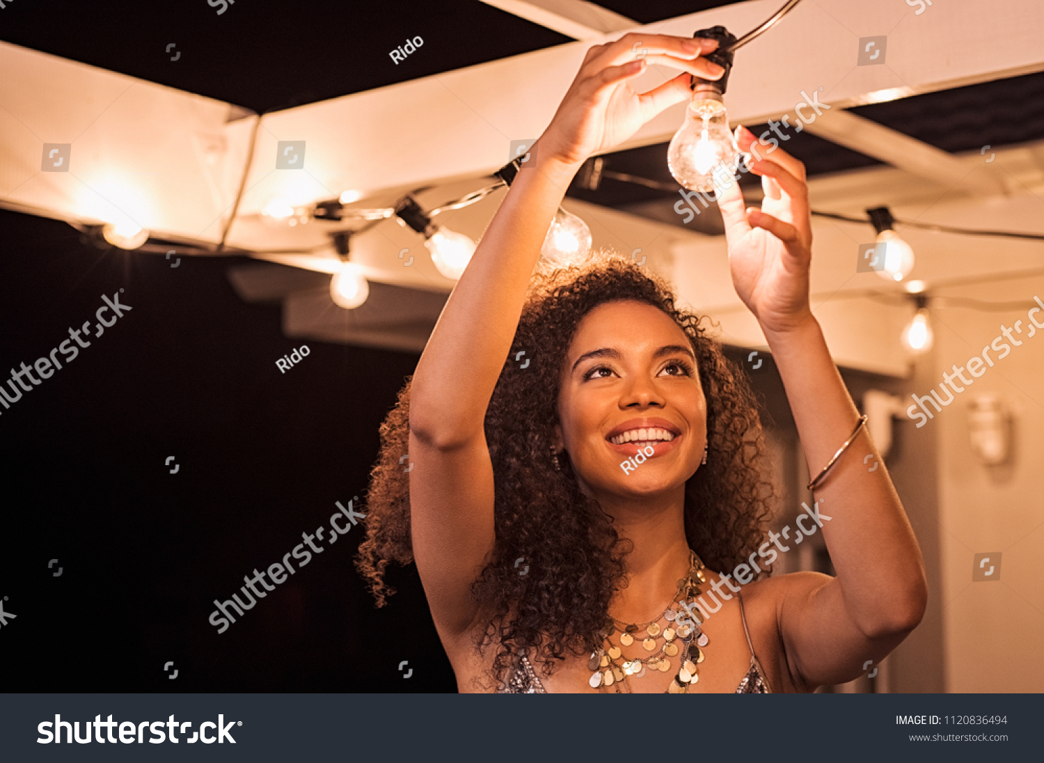 Cheerful young woman wearing party dress changing bulb light in patio. Happy smiling girl making preparation of party by adding lights outdoor. Happy beautiful woman enjoying fixing bulbs in backyard. #1120836494