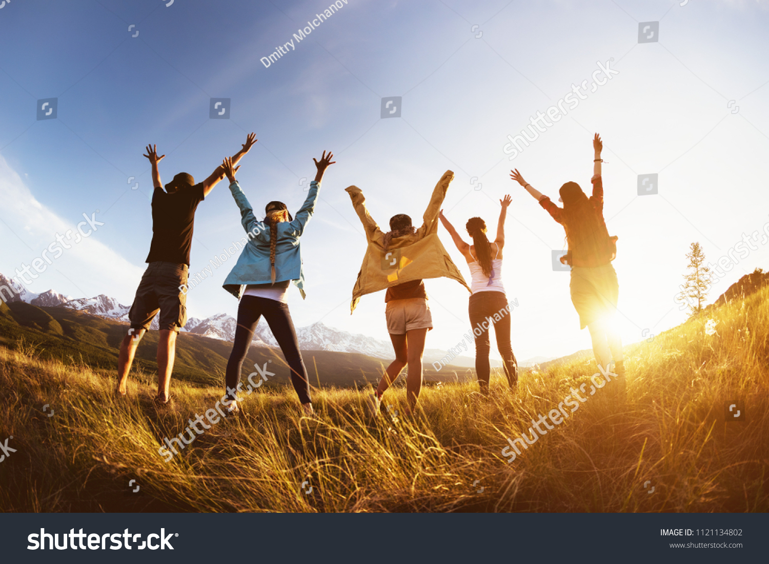 Group of happy friends stands against mountains sunset with raised arms and having fun #1121134802