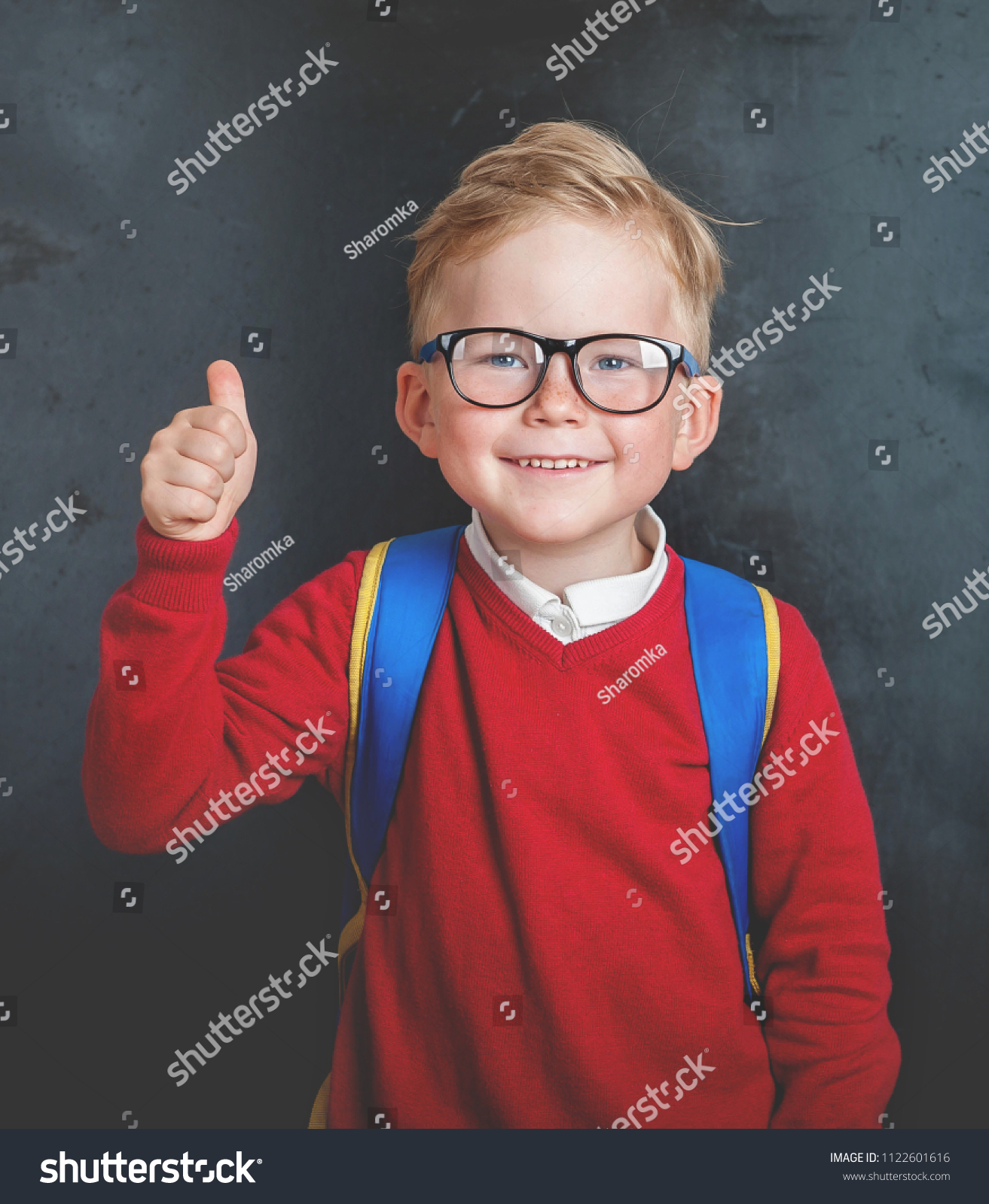 Back to school. Happy little boy in glasses with thumb up and backpack against blackboard.  #1122601616