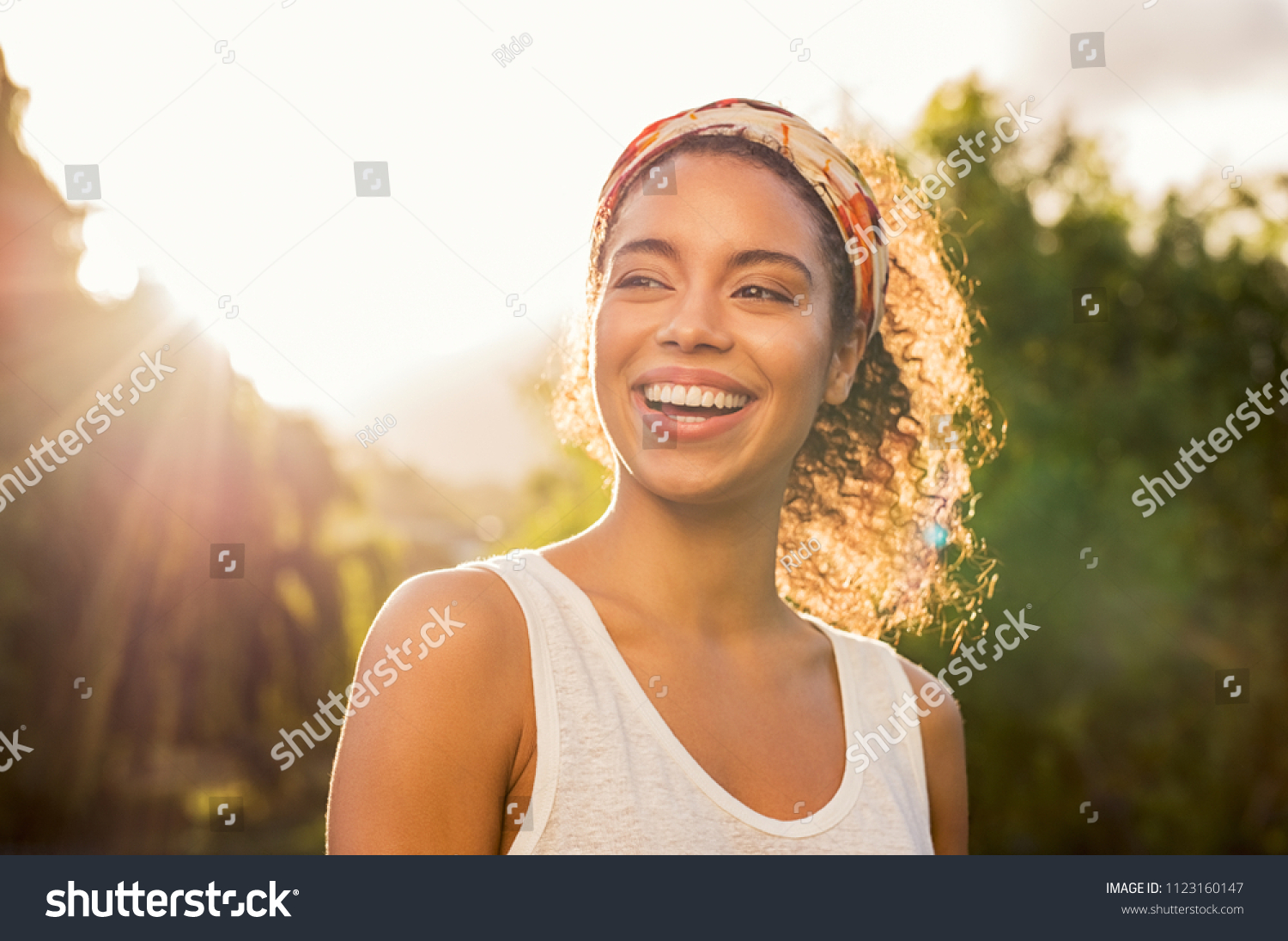 Portrait of beautiful african american woman smiling and looking away at park during sunset. Outdoor portrait of a smiling black girl. Happy cheerful girl laughing at park with colored hair band. #1123160147