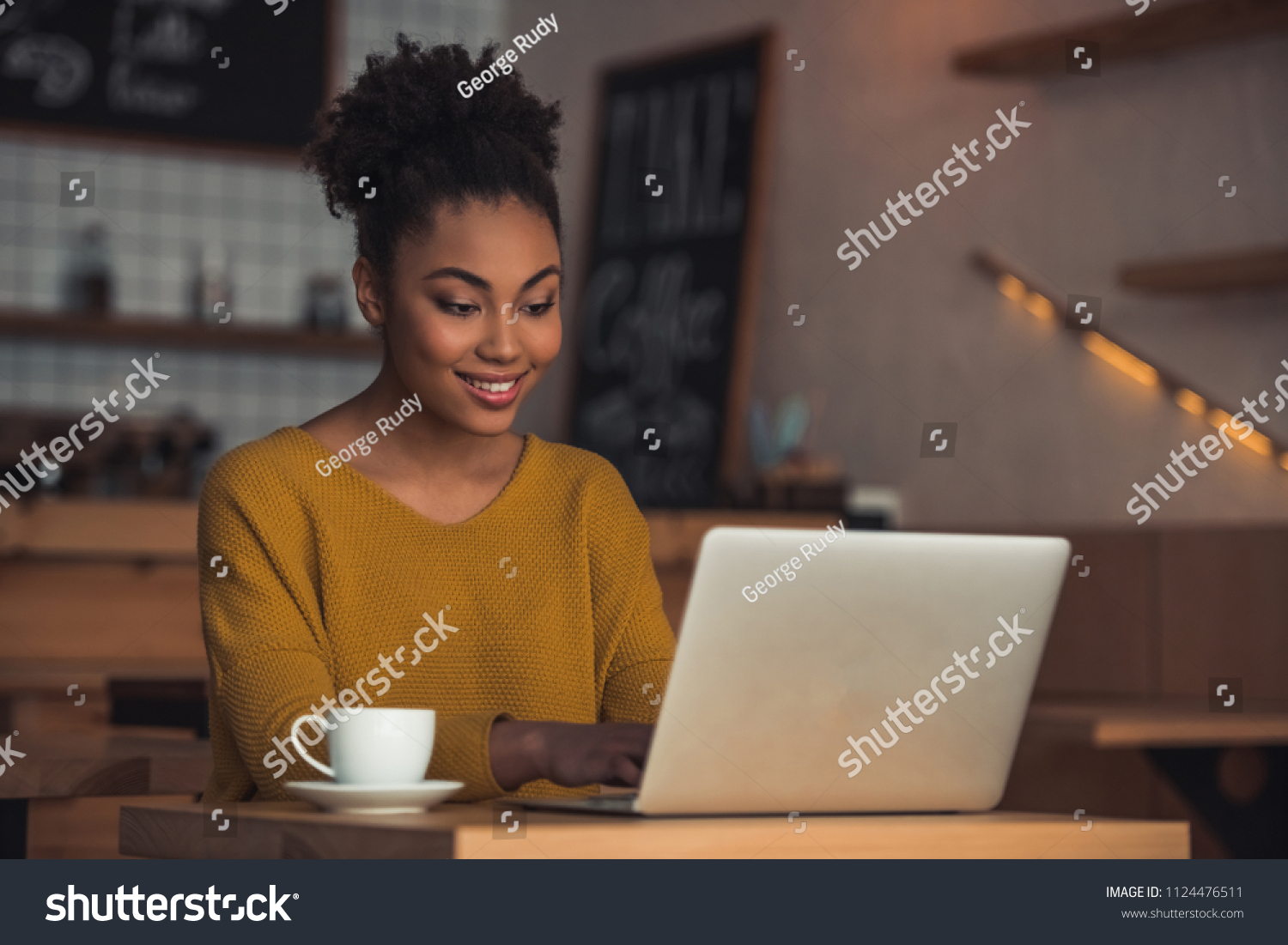Beautiful Afro American girl in casual clothes is using a laptop and smiling while sitting in cafe #1124476511