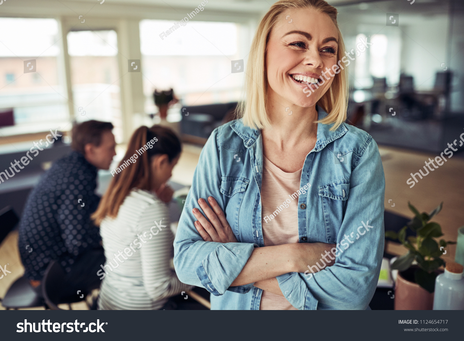 Laughing young businesswoman standing in an office with her arms crossed with coworkers sitting together at a table in the background #1124654717