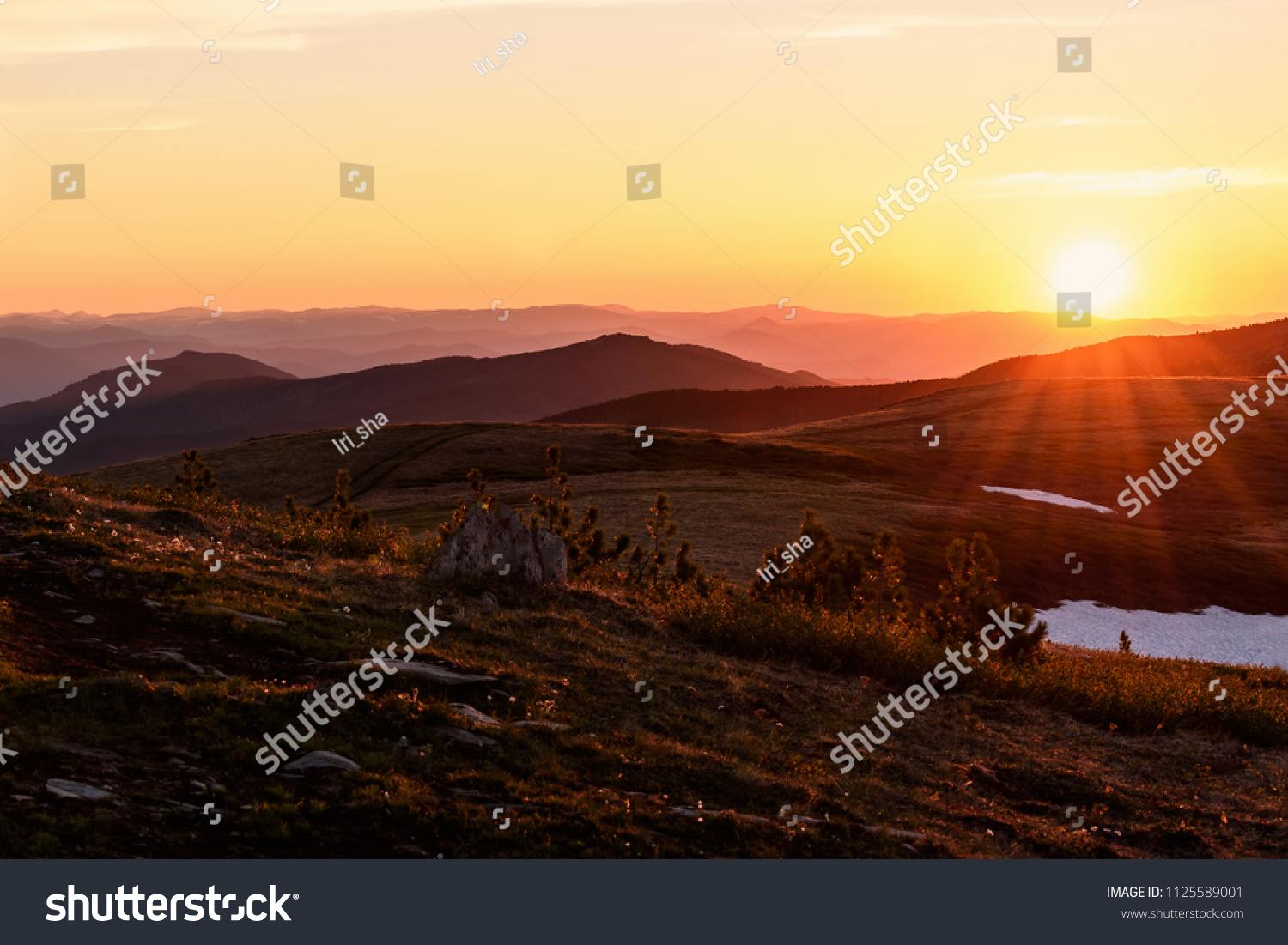 Beautiful sunset with rays over the contours of mountains with stones, small cedars and snow in the foreground #1125589001