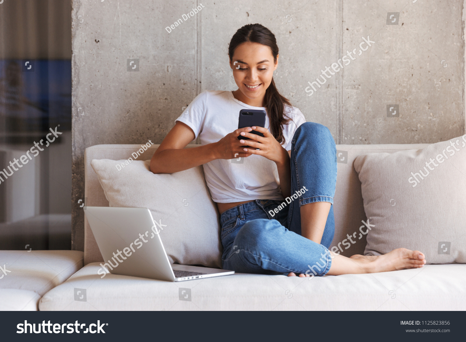 Happy young asian woman using mobile phone while sitting a couch at home with laptop computer #1125823856