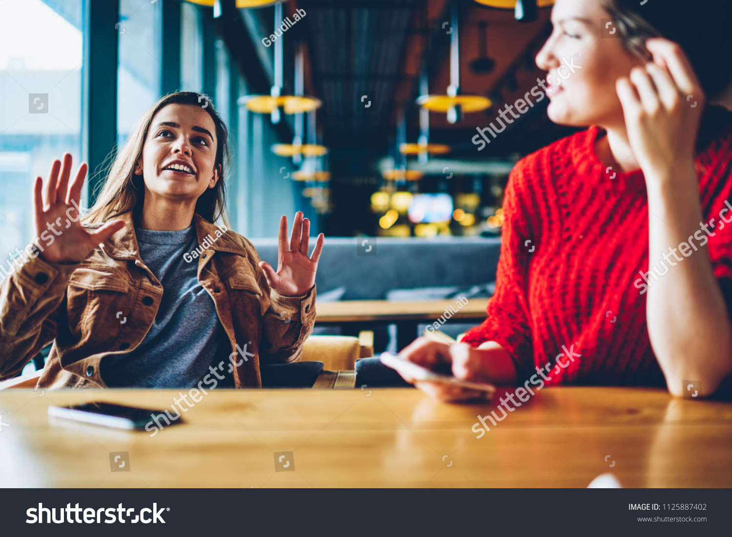 Two happy hipster girls discussing ideas for attracting followers for shared blog in social networks indoors, successful female users communicated at comfortable cafeteria while waiting friend #1125887402