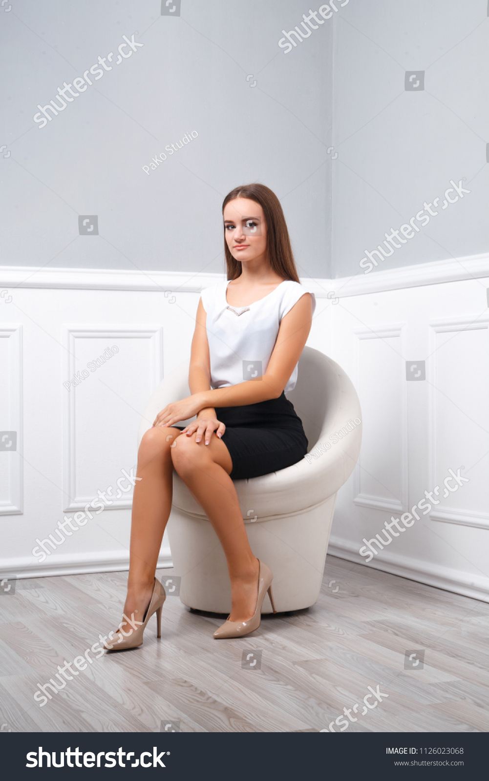 Young beautiful girl sits on a white round armchair in full growth, business style clothes, high heel shoes, leg on leg, looks at the camera. A series of photos #1126023068