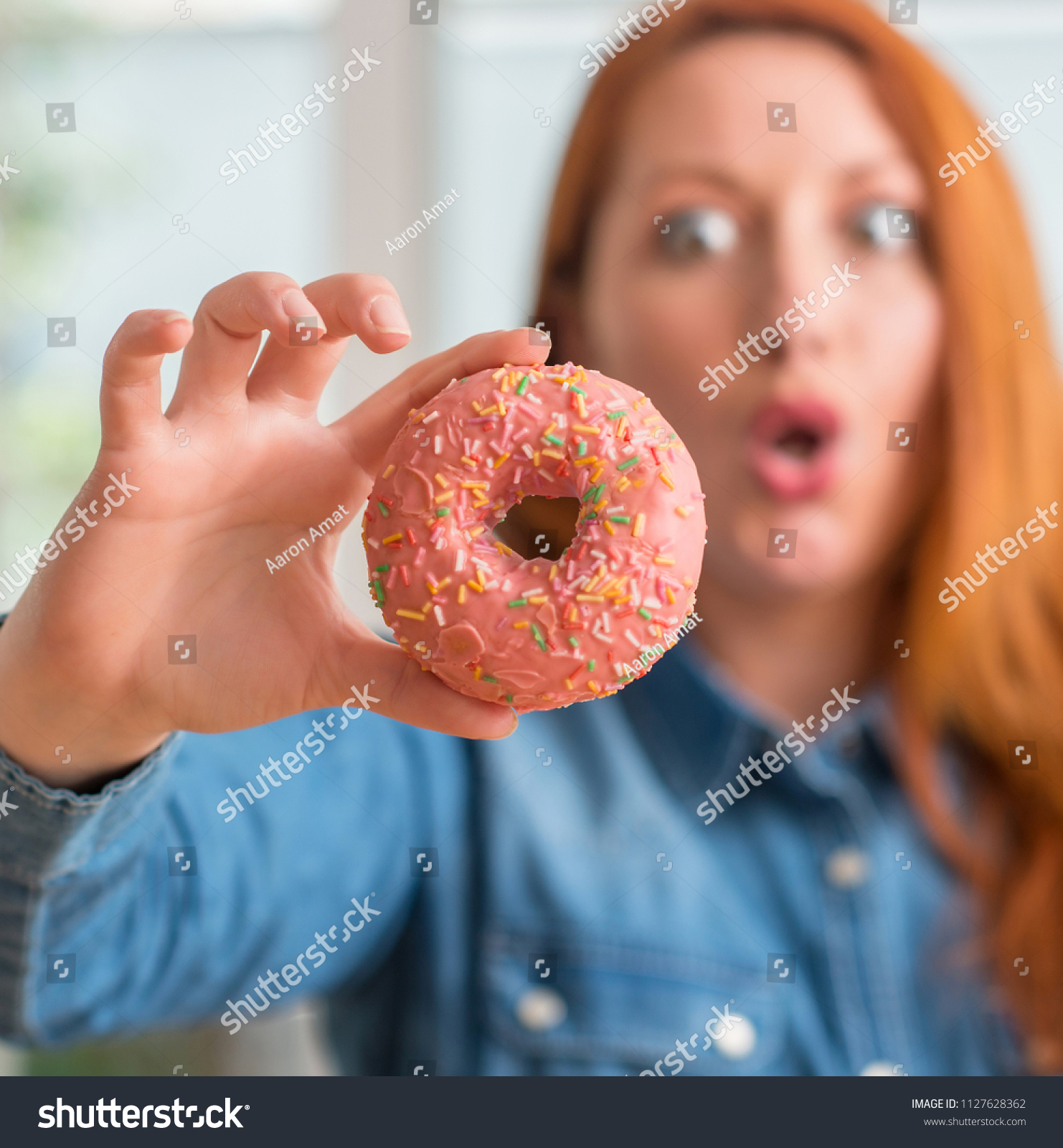 Redhead woman holding donut at home scared in shock with a surprise face, afraid and excited with fear expression #1127628362