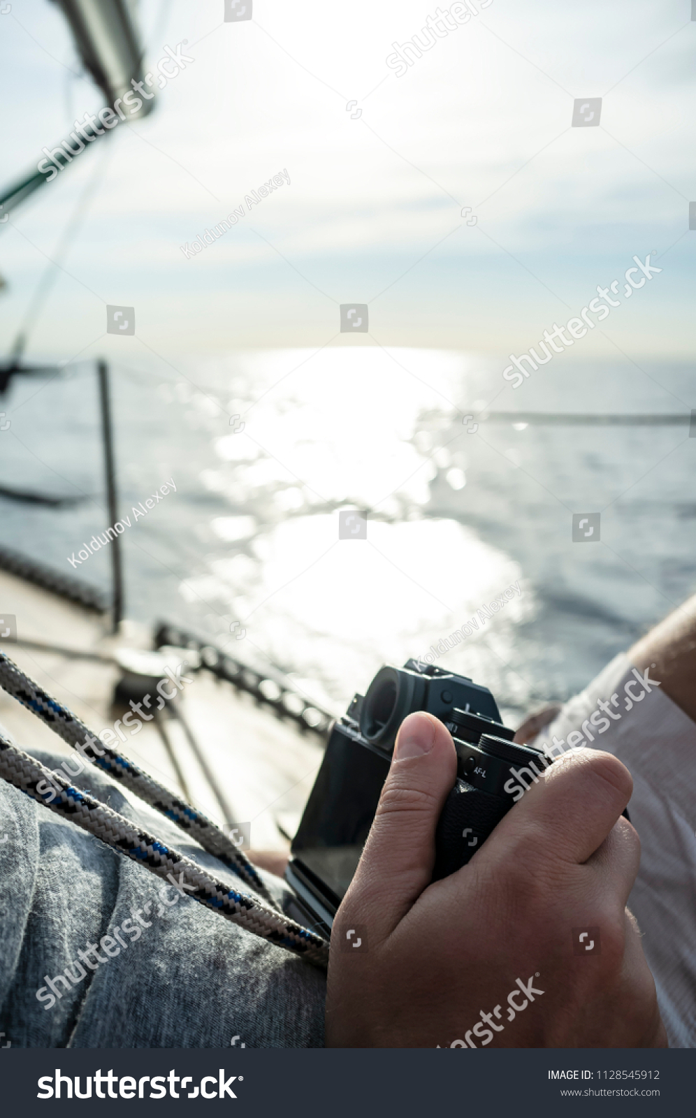 A man with a camera in his hand relaxed sitting on the deck of a yacht, on a blurred background of the sea, sky and sunlight, a warm summer day. #1128545912