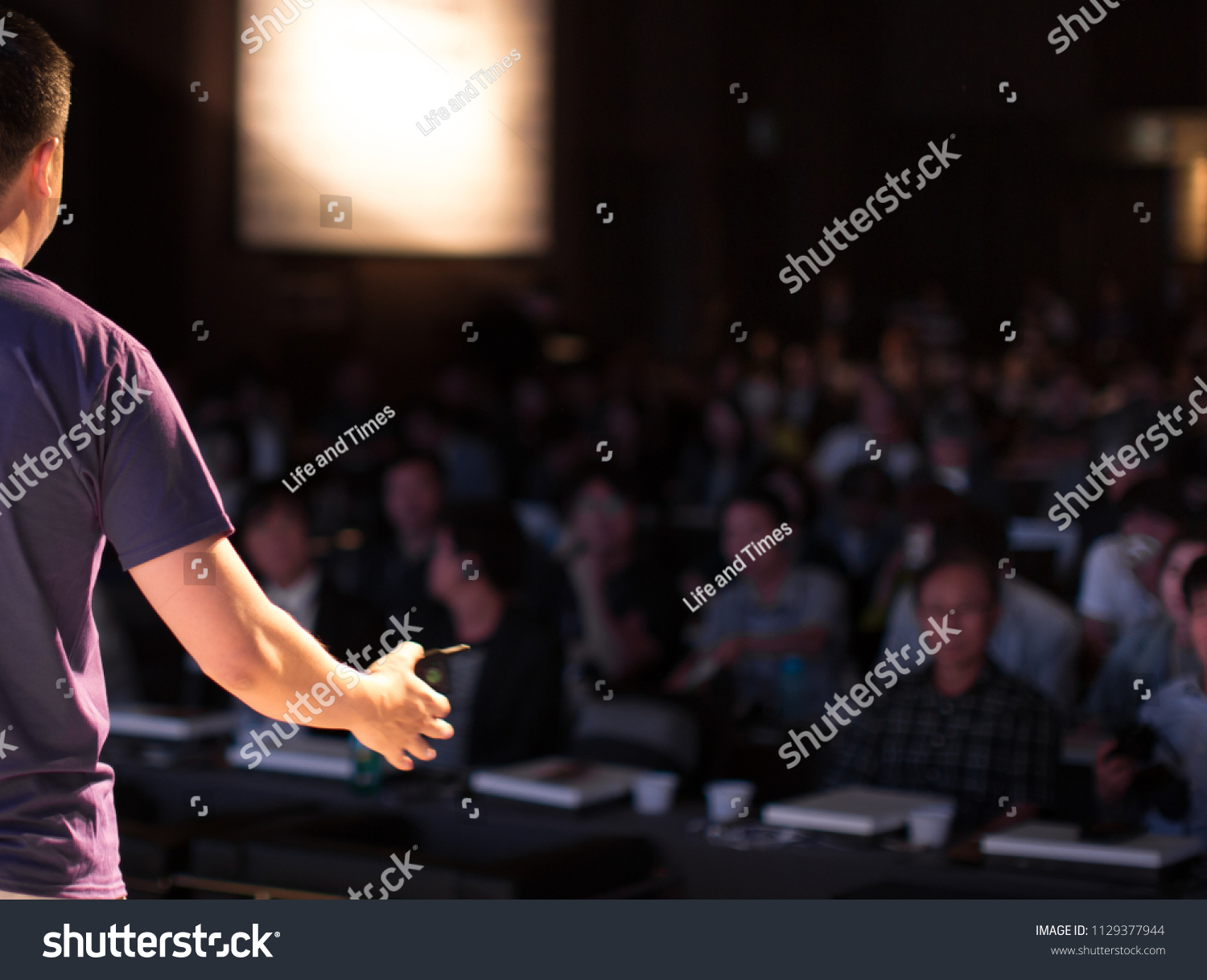 Presenter Speaking to Audience People in Conference Hall Auditorium. Presentation Stage. Blurred De-focused Unidentifiable Audience and Presenter. Technology. Casual Attire Presenter. #1129377944