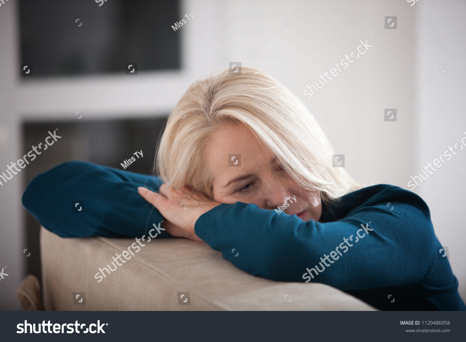 Sad depressed woman at home sitting on the couch, looking down and touching her forehead, loneliness and pain concept #1129486958