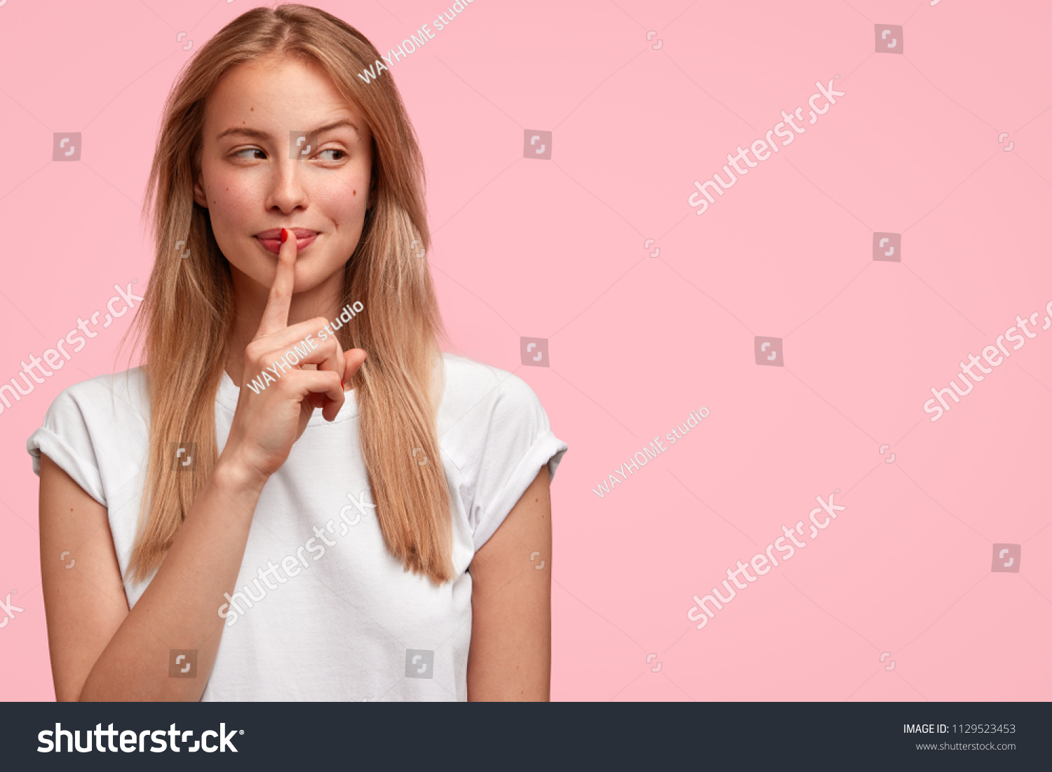 Indoor shot of beautiful female looks mysteriously aside, has intriguing look, asks to be quiet, dressed in casual clothes, stands against pink background with blank copy space for your advertisement #1129523453