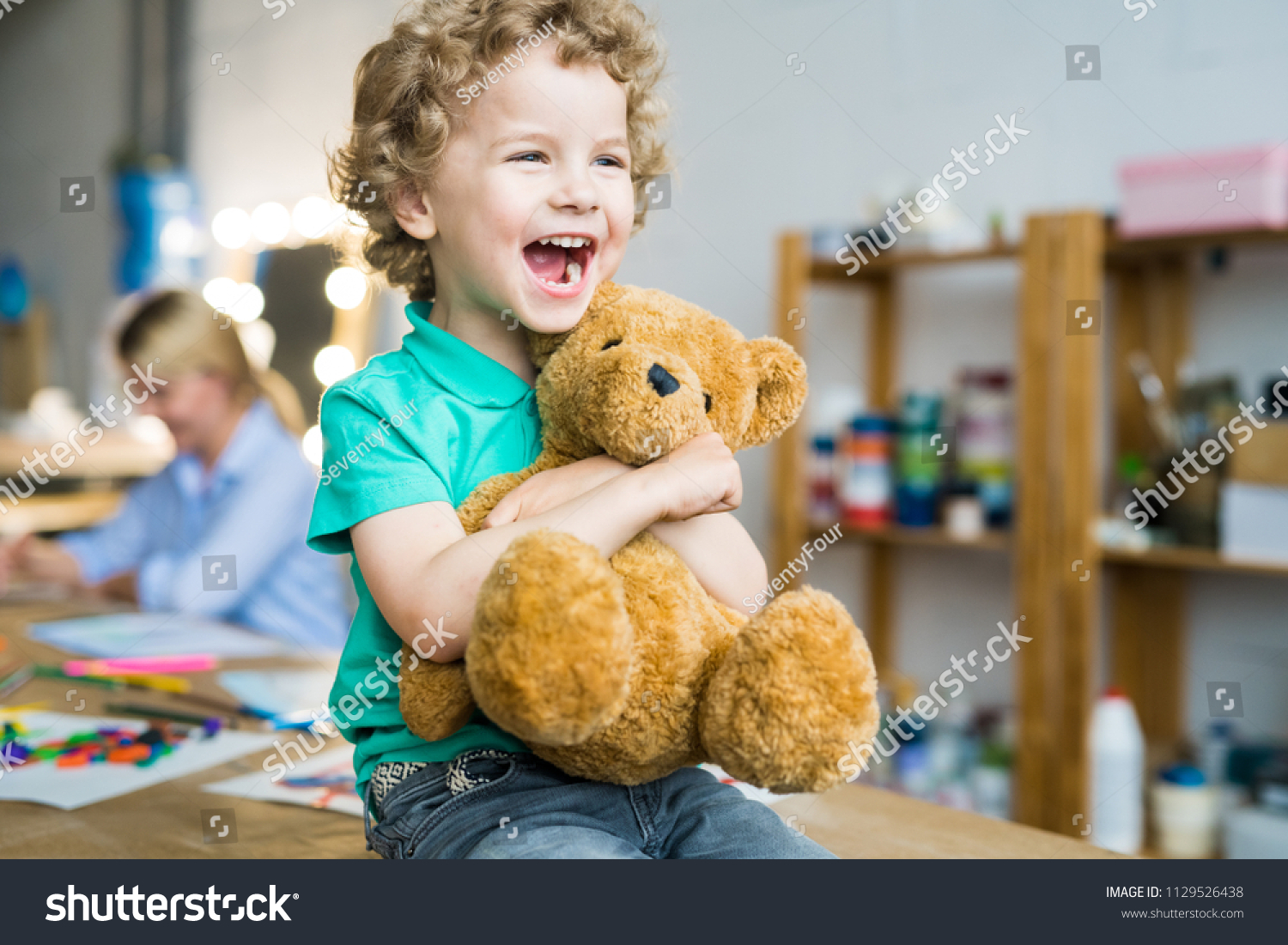 Warm toned portrait of happy curly haired kid laughing cheerfully and hugging teddy bear toy, copy space #1129526438