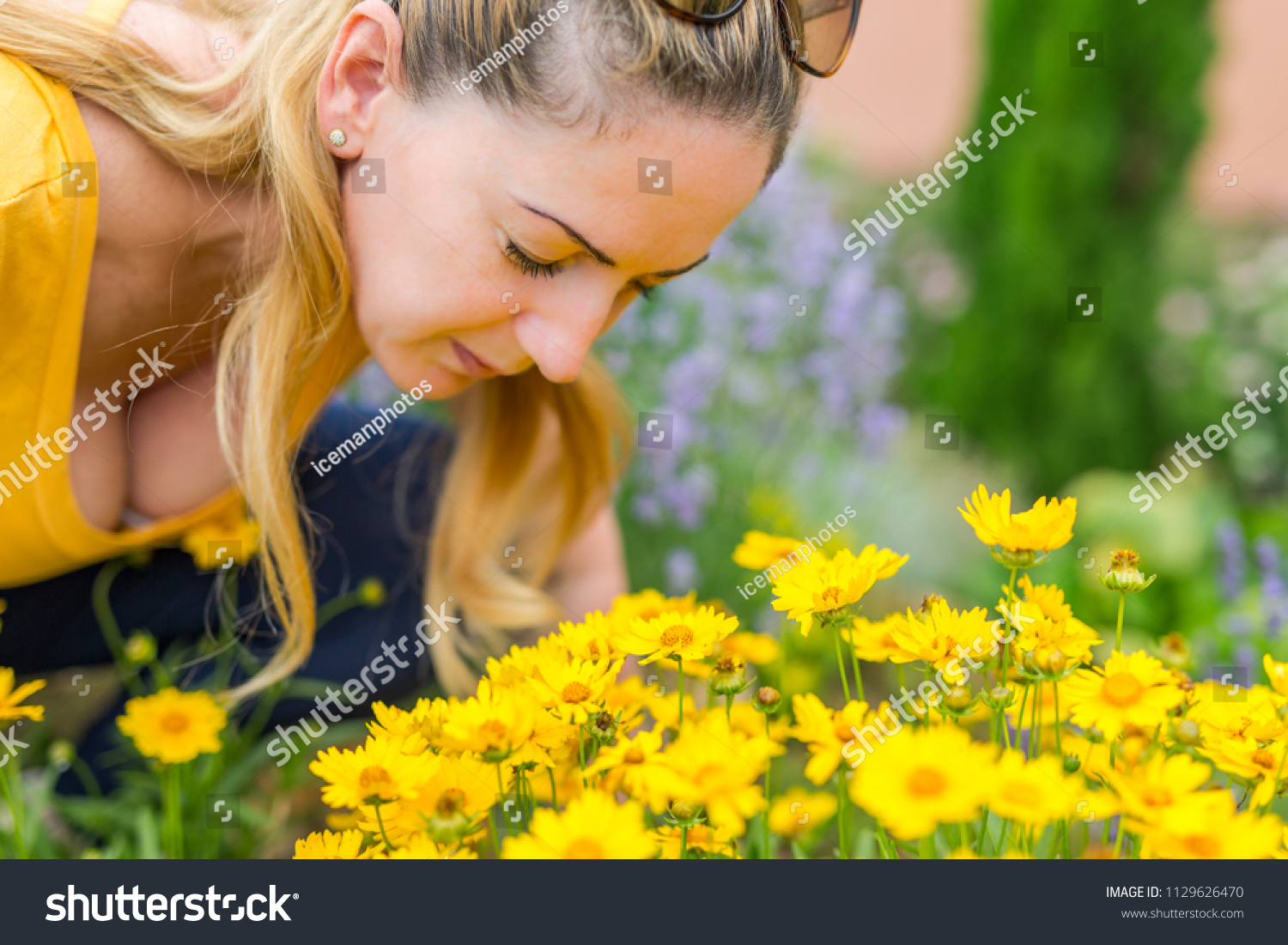 Beautiful blonde woman in yellow top smelling summer flowers in a garden background #1129626470