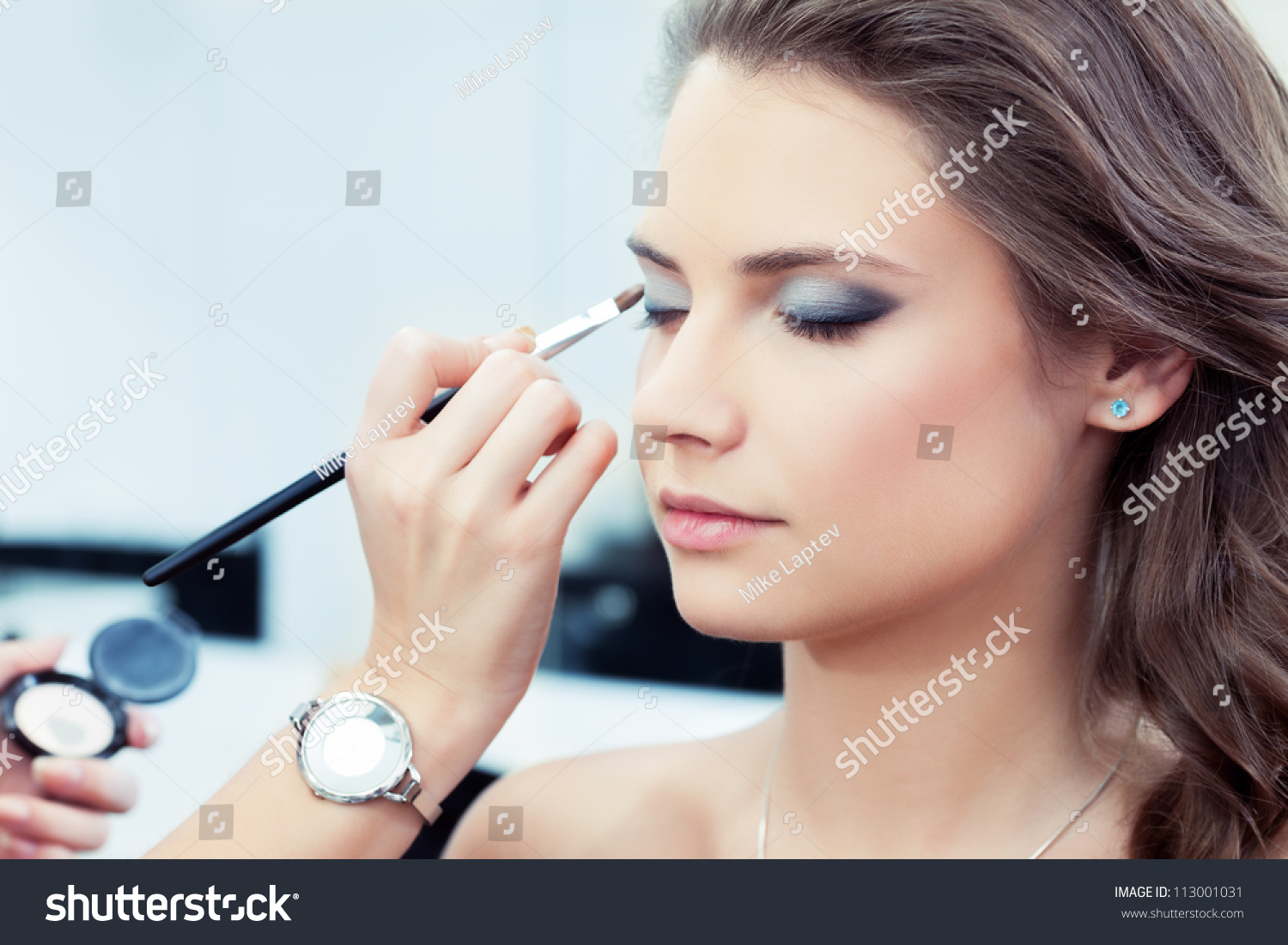 Make-up artist applying bright base color eyeshadow on model's eye and holding a shell with eyeshadow on background, close up #113001031