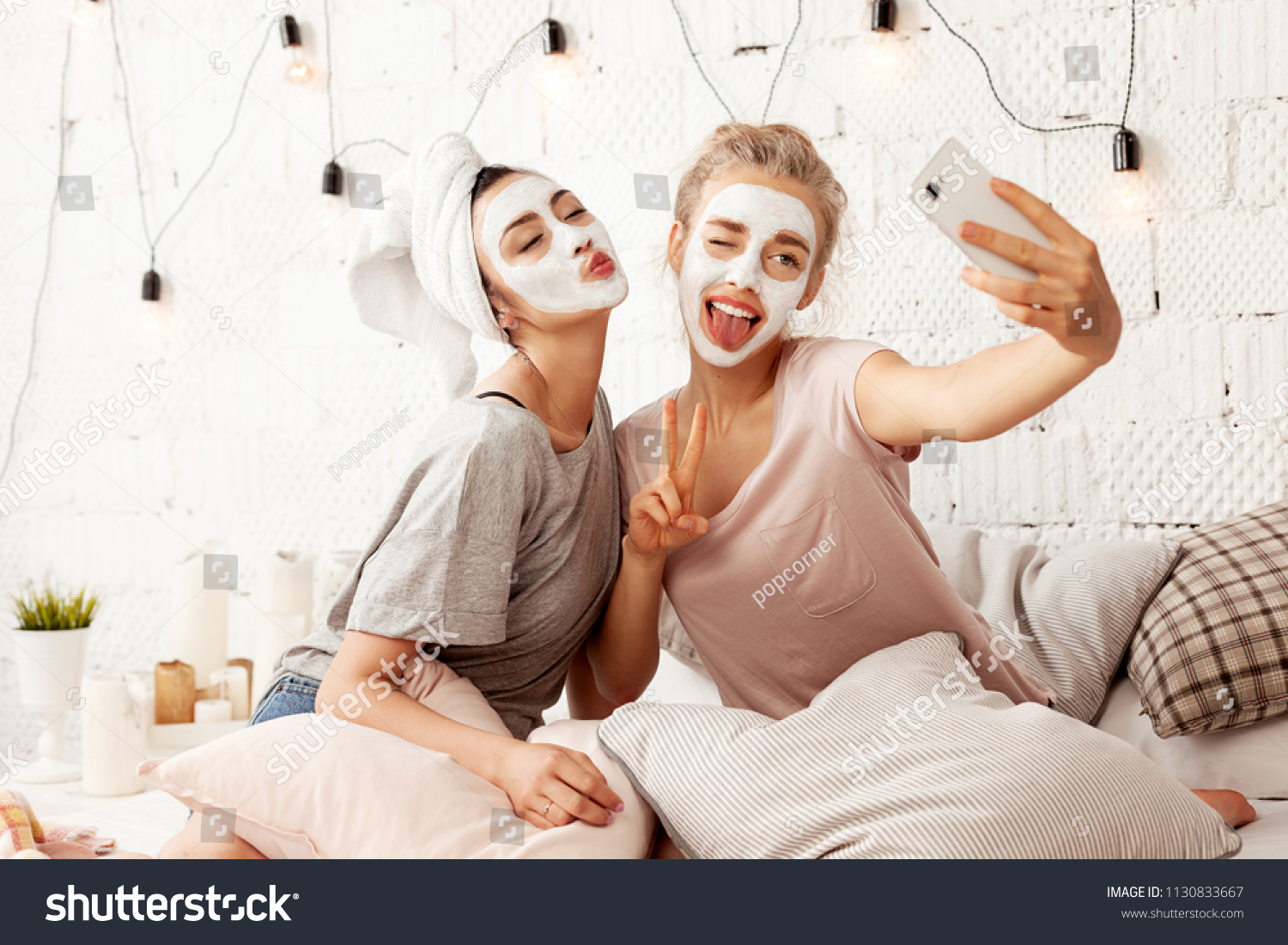 Portrait of cute young women sending kisses smiling sweetly and making amusing faces for picture. Cheerful lovely friends taking selfie at home with facial cosmetic mask. Skincare concept #1130833667