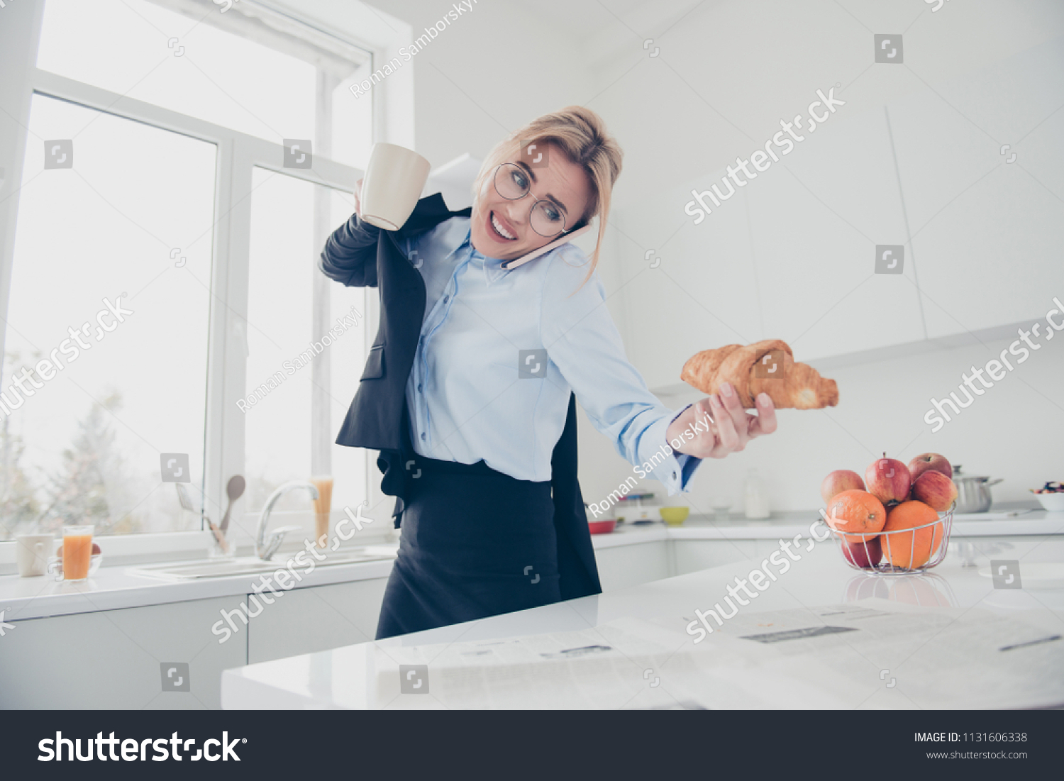 Adorable busy attractive charming beautiful smiling lady office executive worker wearing spectacles in hurry early in the morning talking on the phone having a drink and croissant in kitchen #1131606338