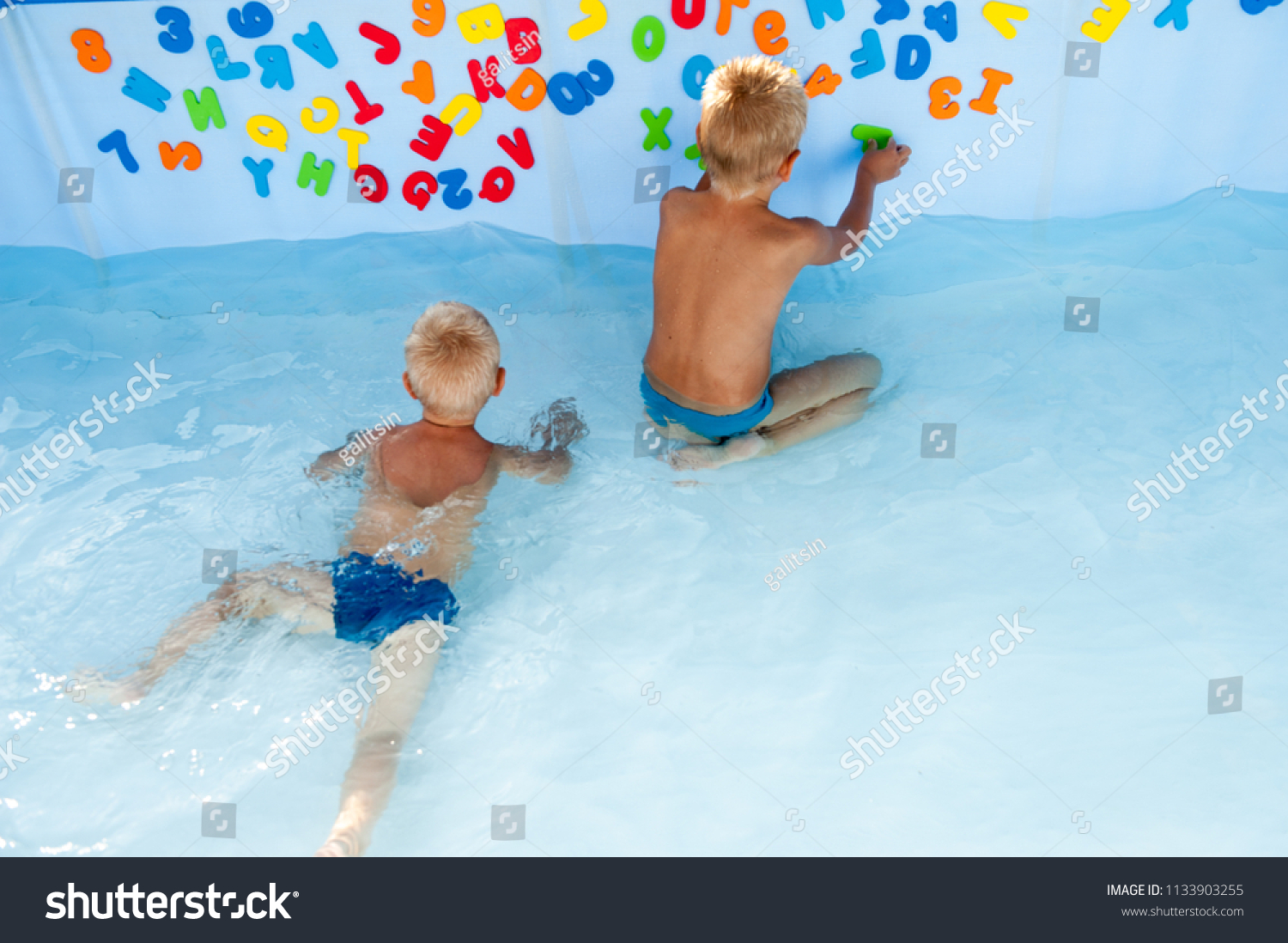 Two happy children play in swimming pool with plastic letters for bathroom. Brothers are happy together in warm water on sunny summer day. Concept of teaching games for preschoolers. Copy space text #1133903255