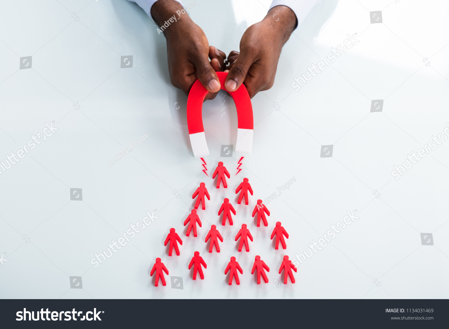 Close-up Of A Human Hand Attracting Red Human Figures With Horseshoe Magnet On White Background #1134031469