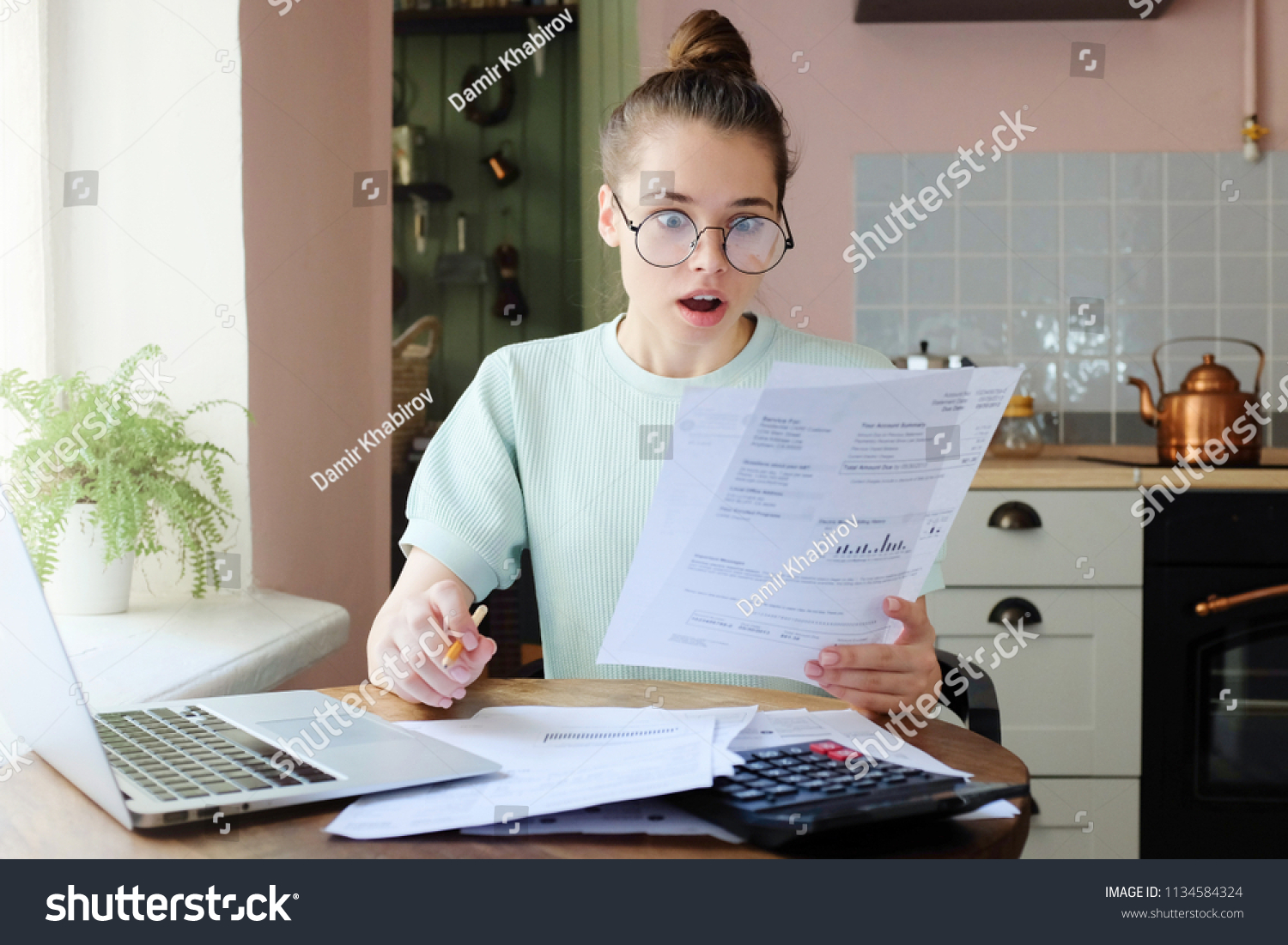 Horizontal closeup of European Caucasian lady spending afternoon at kitchen table, wearing eyeglasses, looking shocked with what she sees in documents she is dealing with, dissatisfied with debts #1134584324