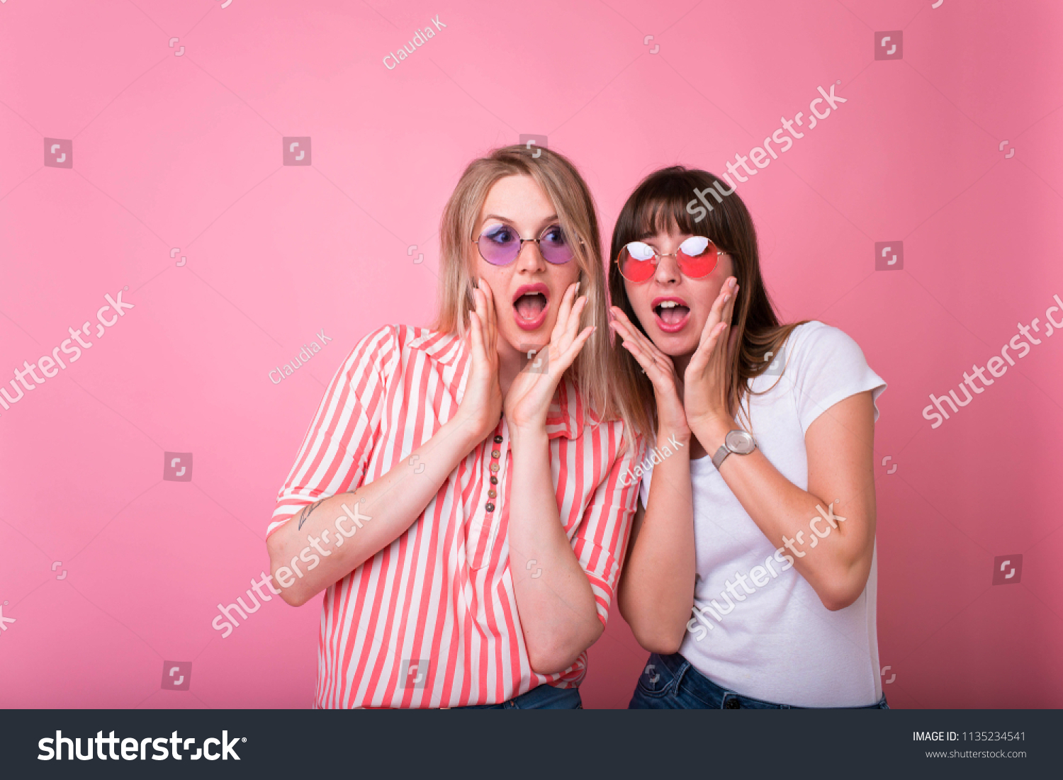 Two cheerful excited young women shouting over pink background.Studio shot of two scared Caucasian female student. raising brows and keeping hands on faces #1135234541