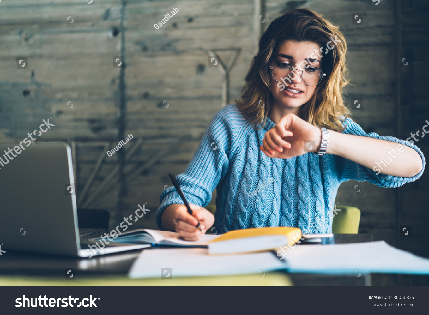 Caucasian hipster girl checking time on wristwatch feeling unhappy and hurrying up on deadline learning and doing homework using literature and laptop at wifi zone, stressed student preparing for exam #1136056829