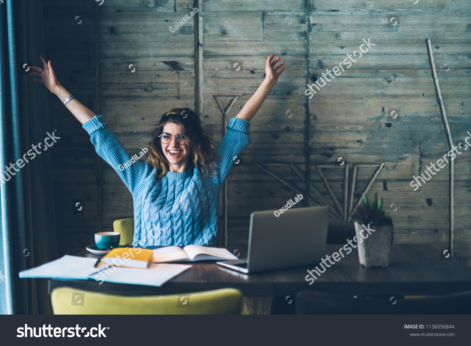 Student girl sitting at table with open notebook and raises hands up while smiling with sincere happiness. 
Woman overjoyed about successfully completed project on computer. Concept of achievement #1136056844