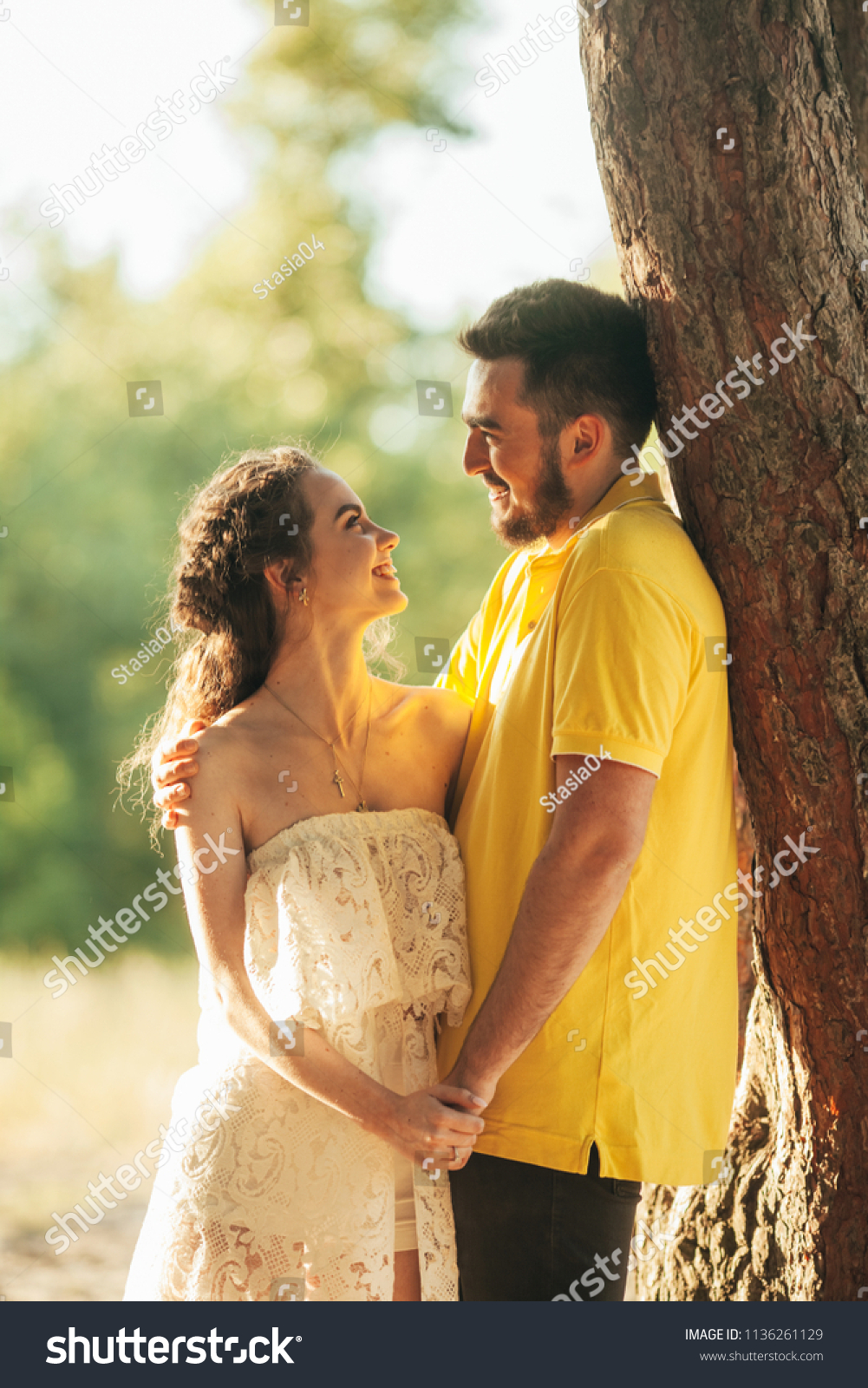 Young enamored couple smiles, hugs and holds hands in forest against background of tree. #1136261129