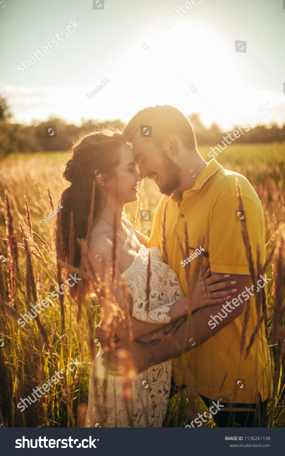 Young enamored couple smiles and hugs at meadow against background of grass and spikes. Backlit. #1136261138