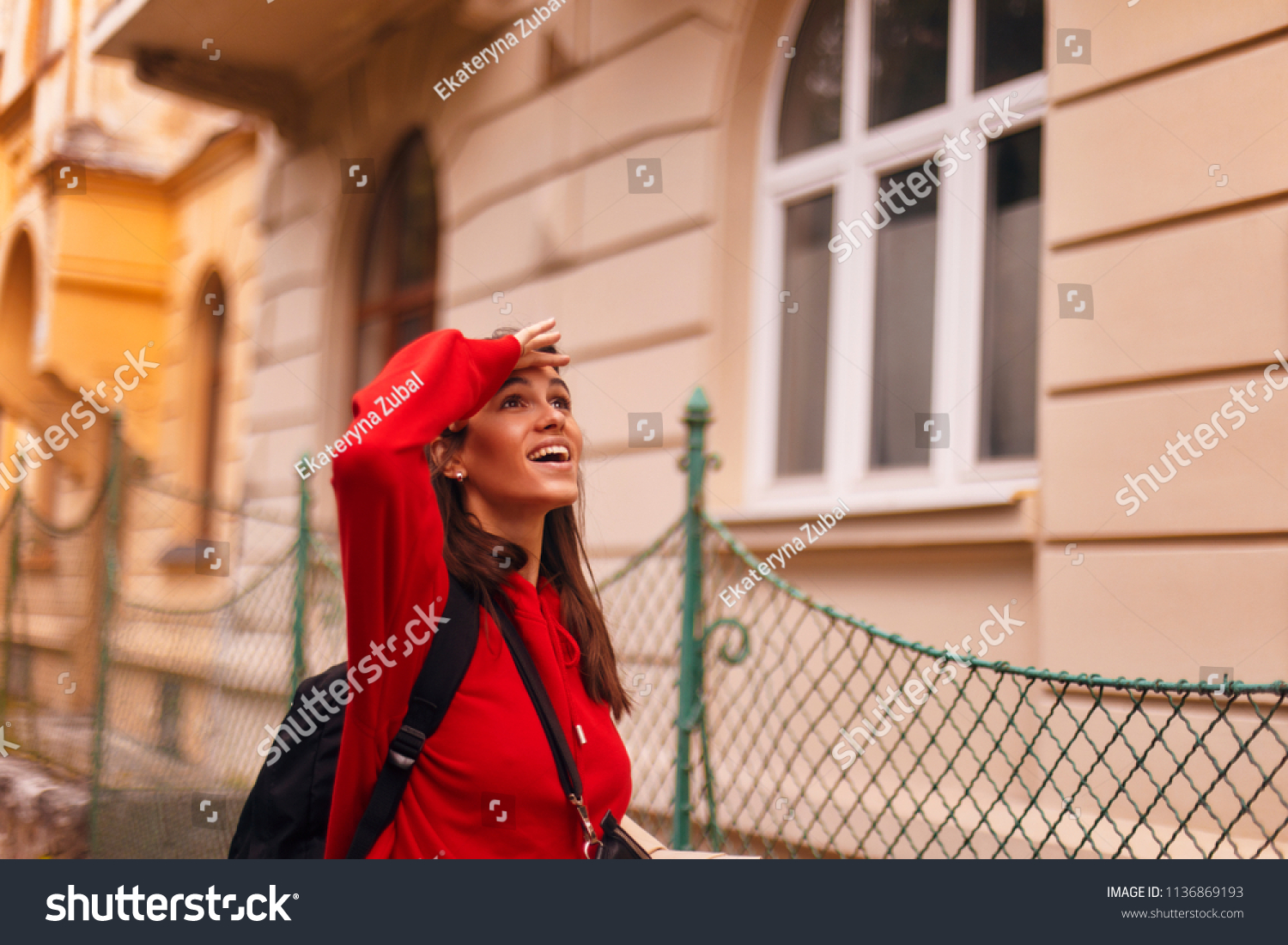 happy tourist woman walking on the street with nice architecture. brunette girl is looking at something. wearing red hoodie and with packed black bag #1136869193