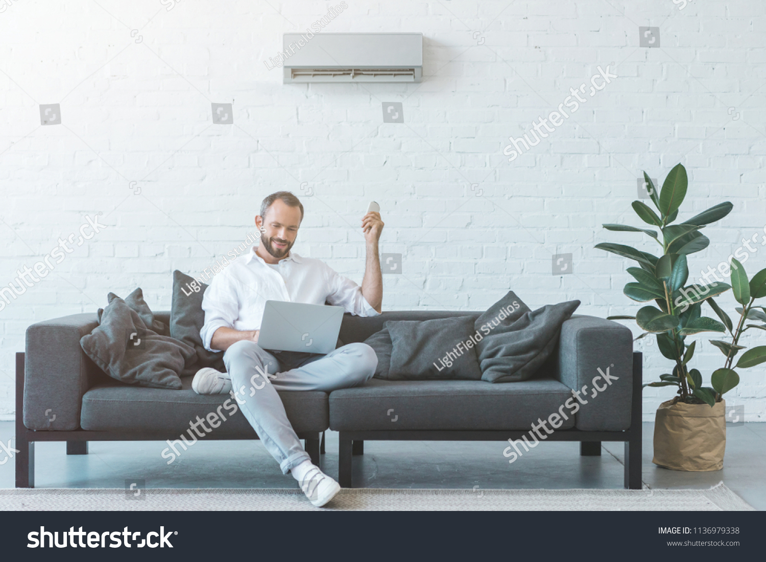man turning on air conditioner with remote control while using laptop on sofa #1136979338