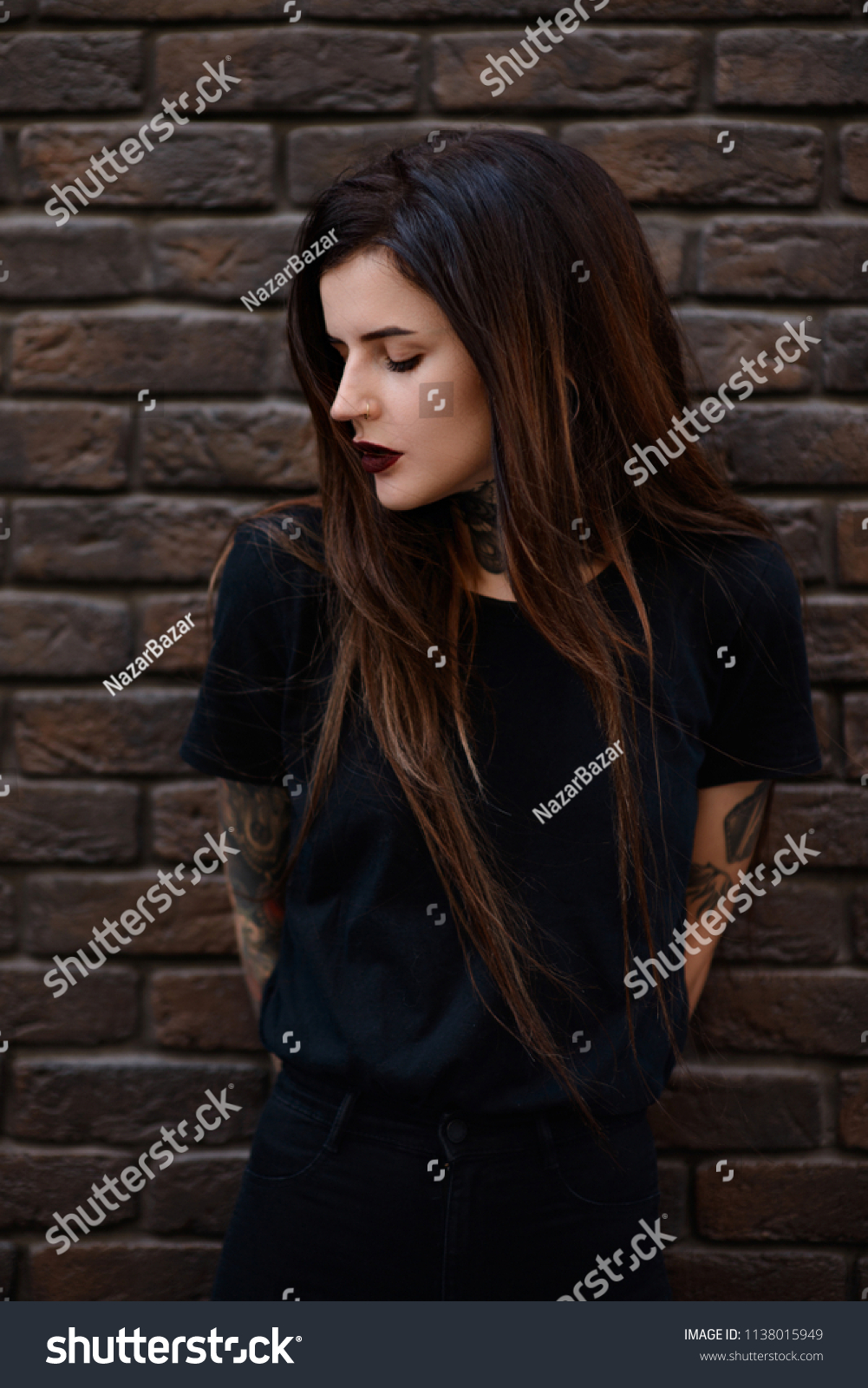 Female model with tattoos, piercing in nose, black lips and long brunette hair. Beautiful slim girl with tattoos wearing black t shirt and jeans standing against the stone wall or brick fence. #1138015949