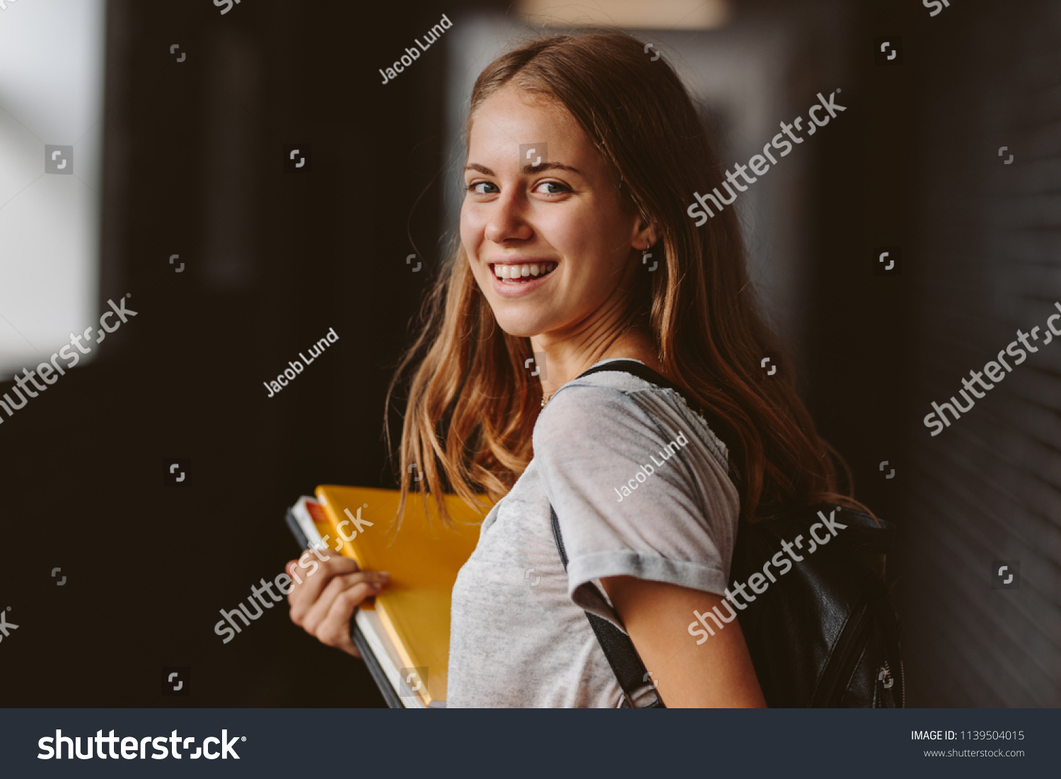 Rear view of beautiful girl walking through university hallway looking back and smiling. female student going for the lecture. #1139504015