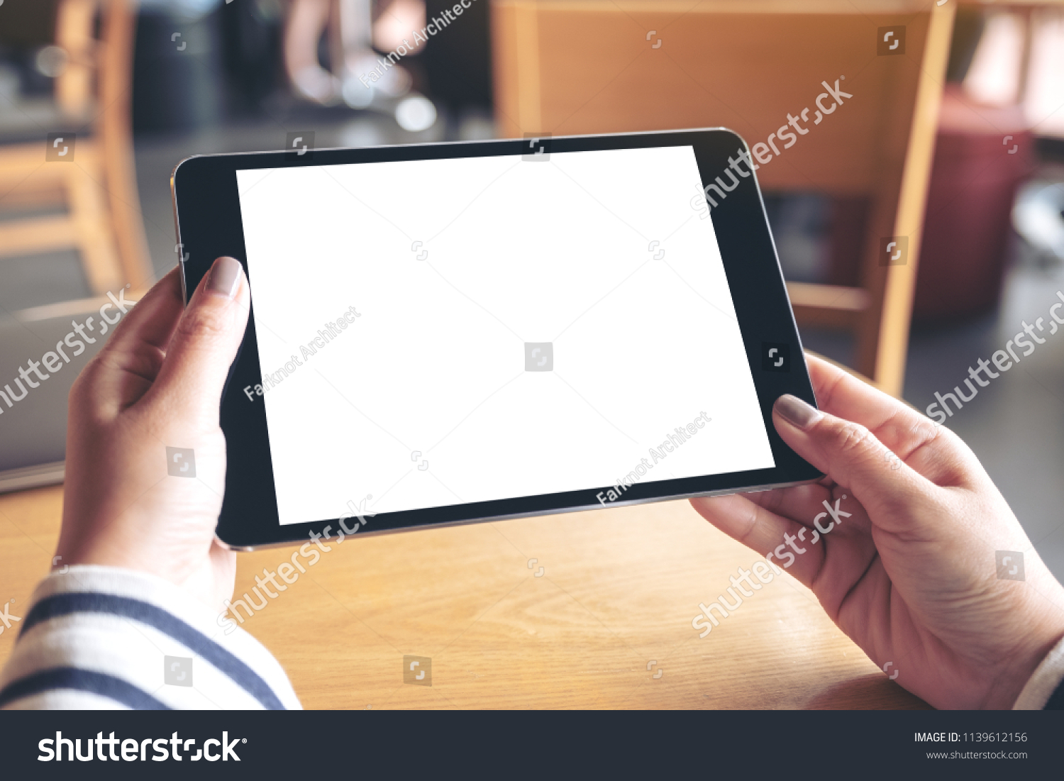 Mockup image of hands holding black tablet pc with blank white desktop screen on wooden table in cafe #1139612156